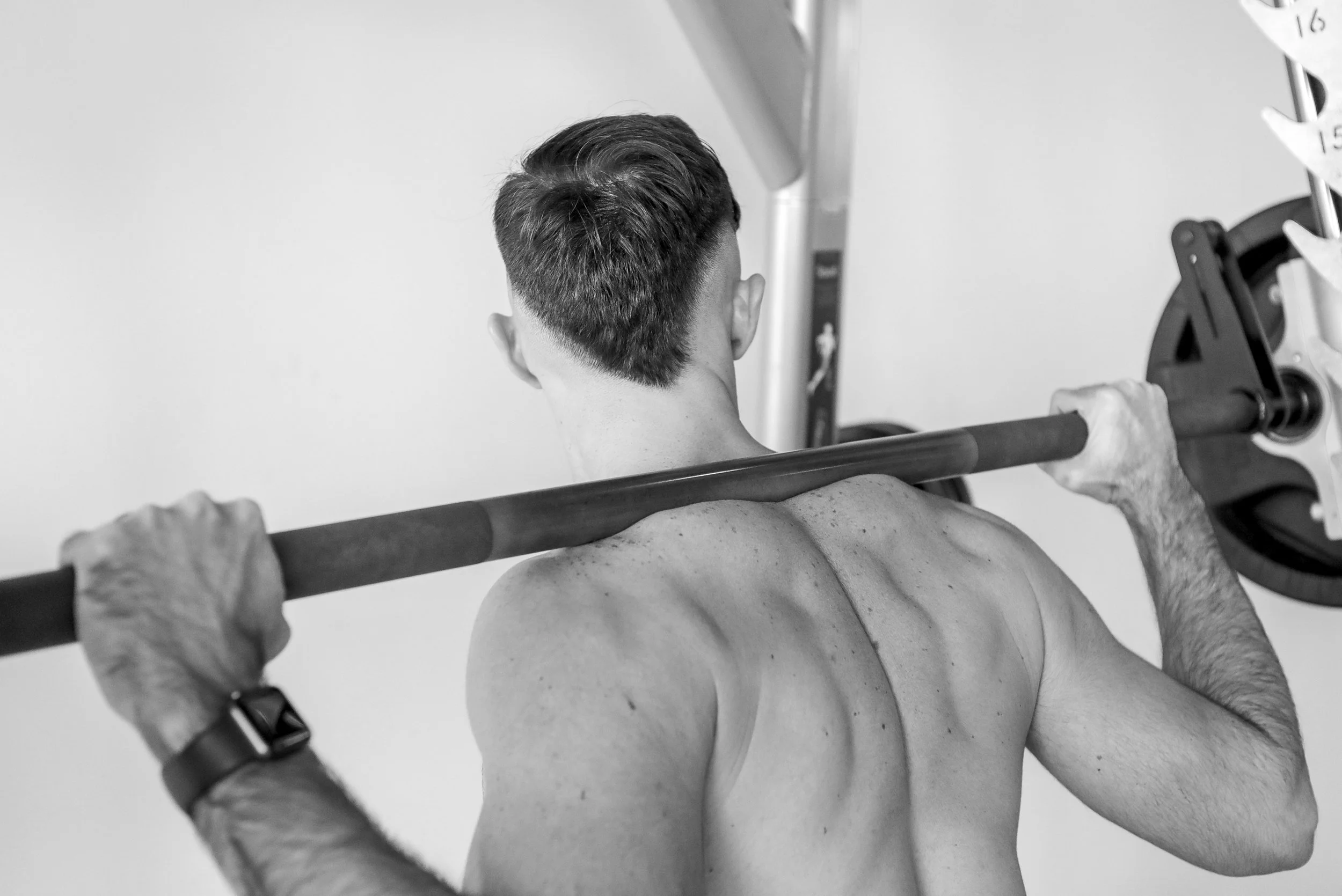 Back view of a shirtless man lifting a barbell during a workout in a gym.