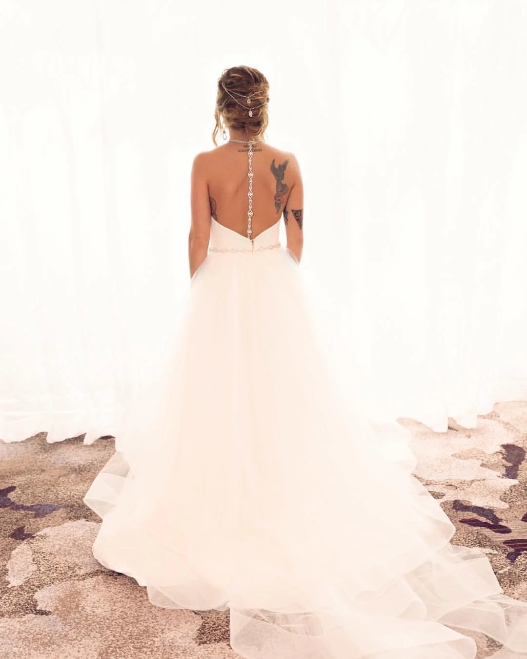 Back view of a woman in a white wedding dress with tattoos on her back and arms, standing in front of white curtains and a patterned carpet.