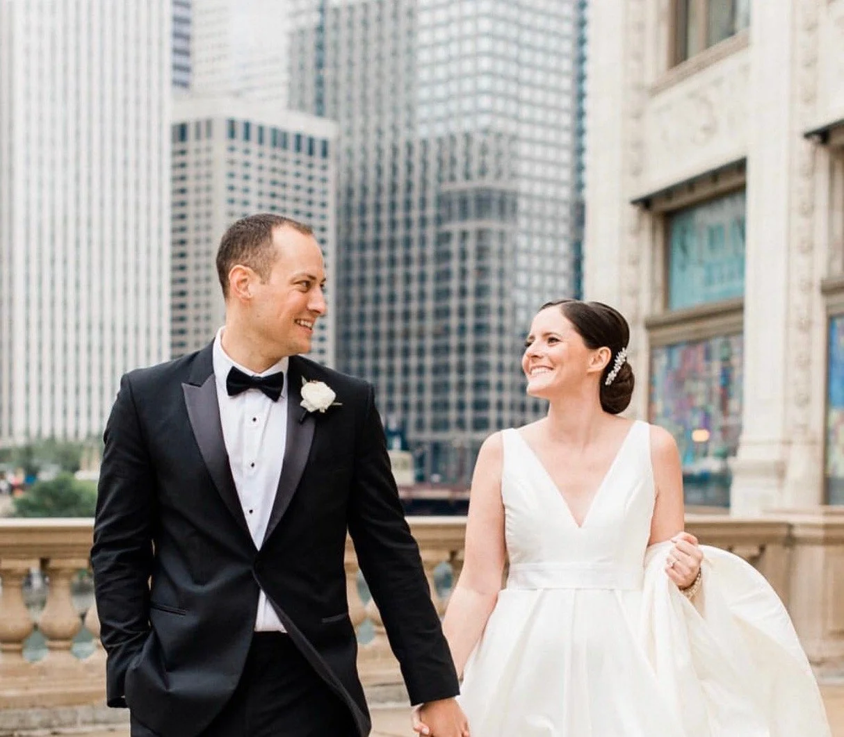 A bride and groom walking hand in hand outdoors in a city, smiling at each other. The groom wears a black tuxedo with a bow tie. The bride wears a white wedding dress with a deep V neckline and is holding part of her dress. Tall buildings are in the 
