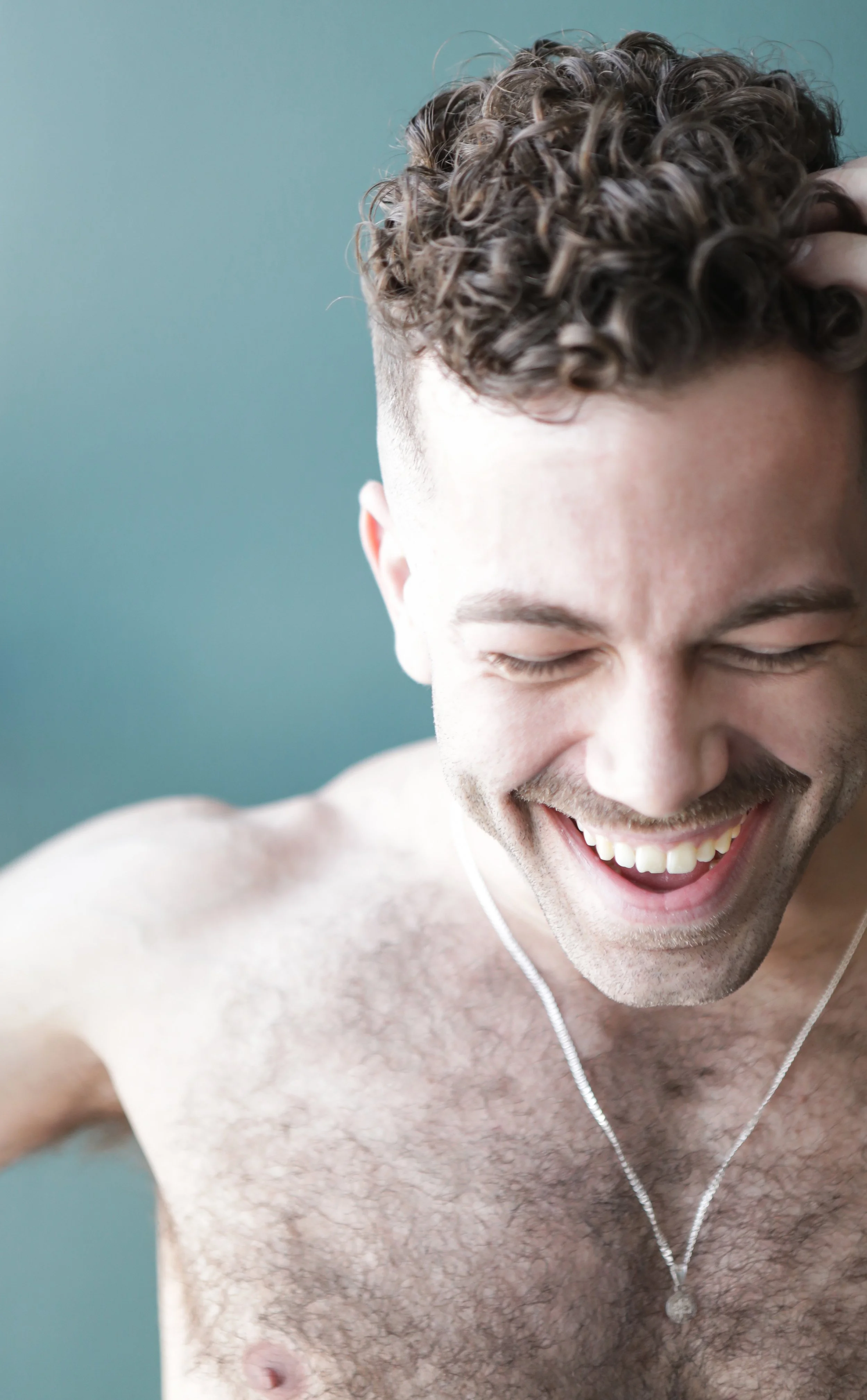 A close-up of a smiling man with curly hair, closed eyes, and visible chest hair, wearing a silver necklace, against a teal background.