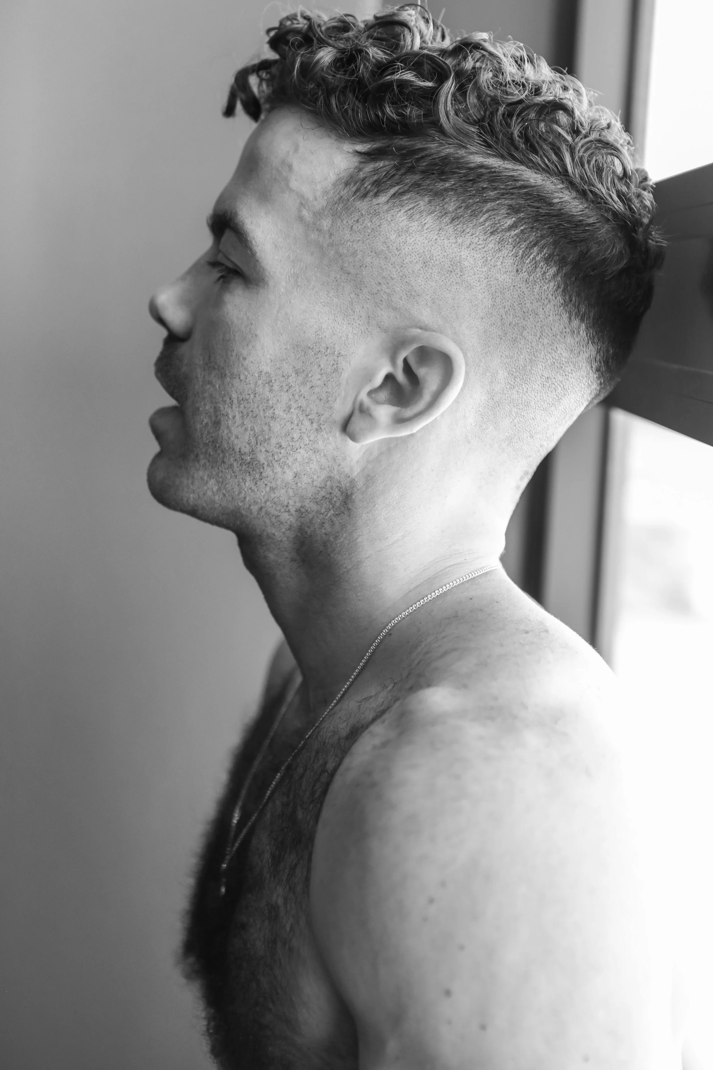 Black and white photo of a young man with curly hair and a fresh fade haircut, seen in profile, shirtless with a necklace, standing by a window.