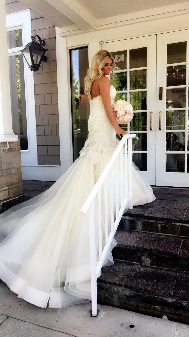 A bride in a white strapless wedding gown with a long train stands on the stairs outside a building holding a bouquet of pink and white roses.