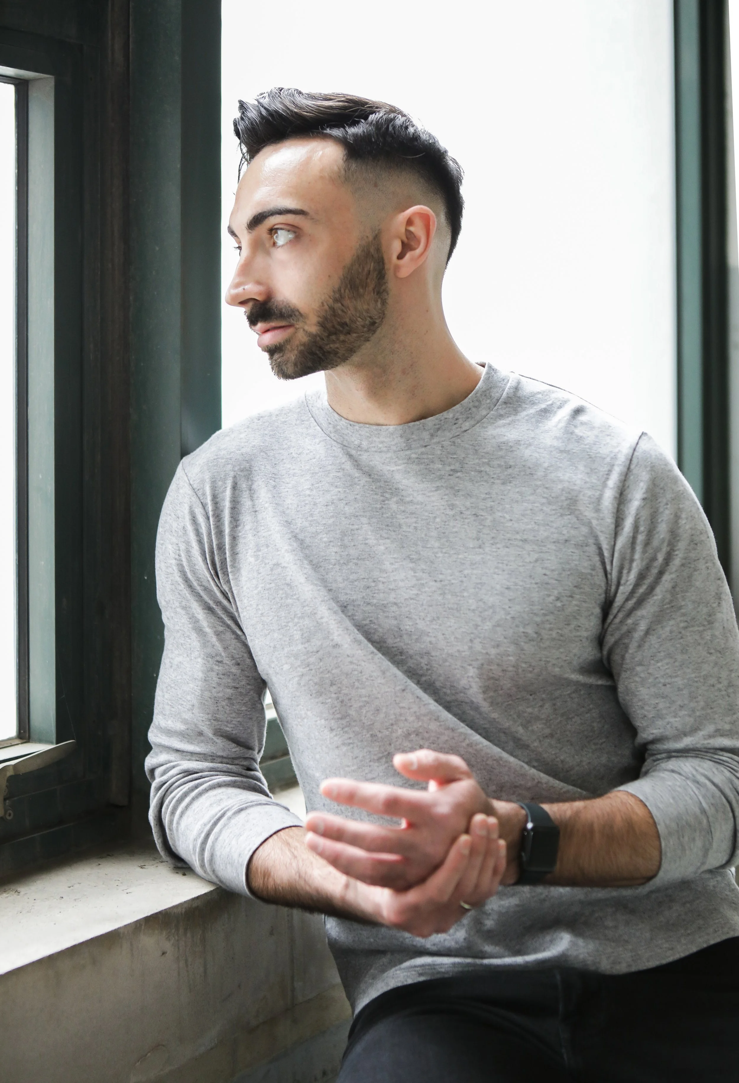A man with dark hair and a beard, wearing a gray shirt and a black watch, sitting by a window and looking to the side.