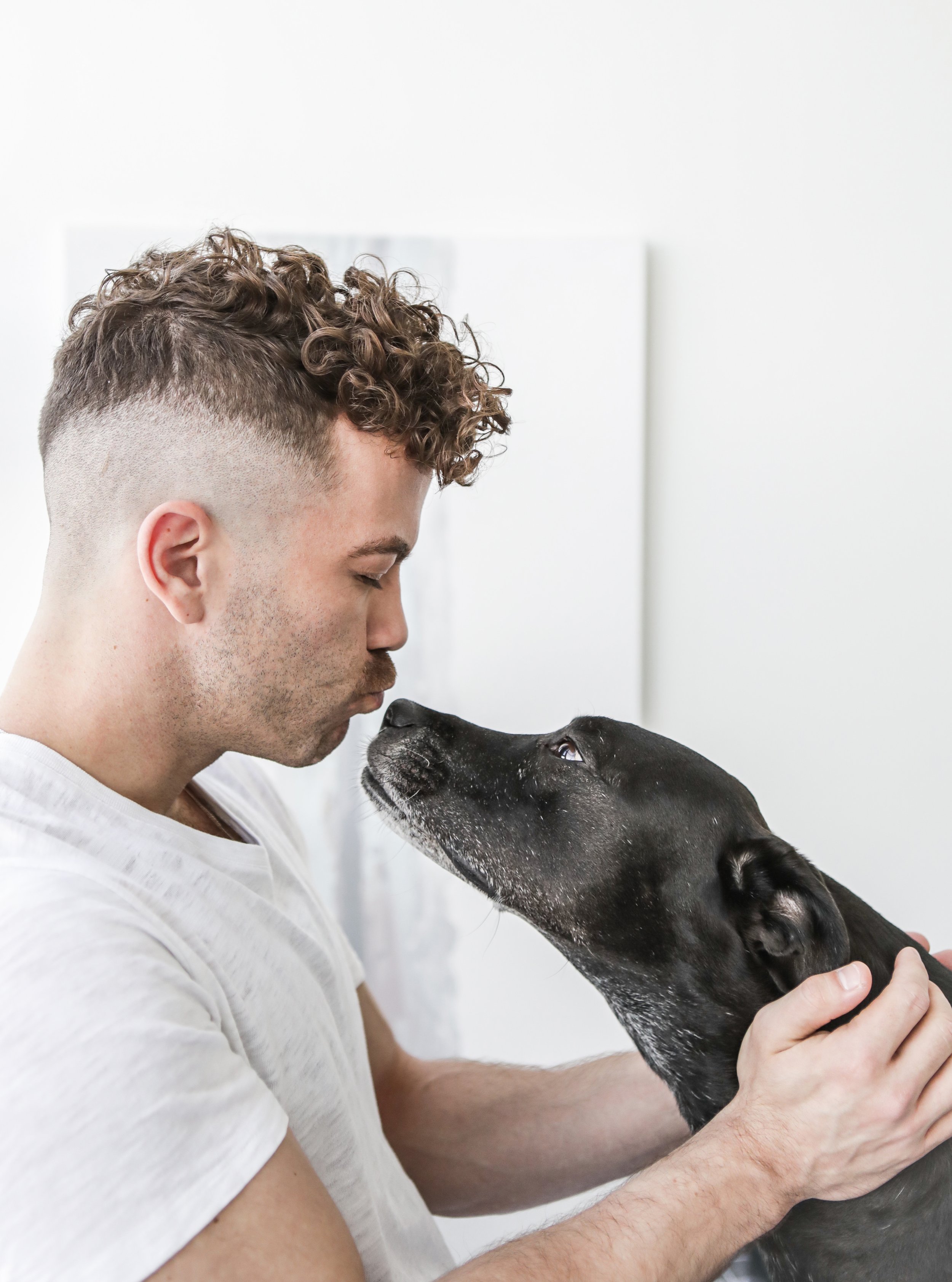 A man with curly hair and a white t-shirt gently holds a black dog, noses touching, inside a bright, minimalistic room.