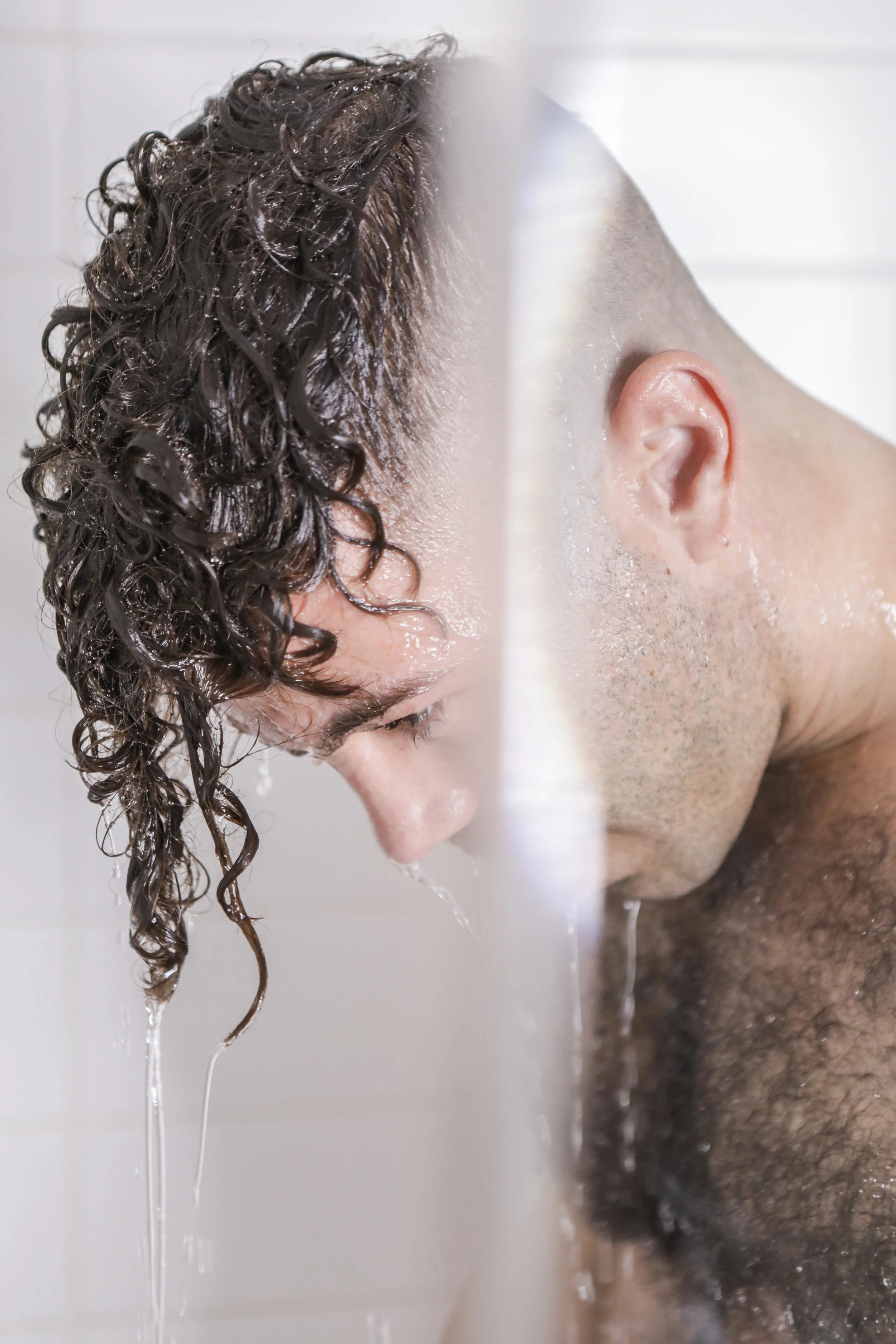 A man with wet curly hair and a beard is washing his face in the shower, with water running down his face and hair.