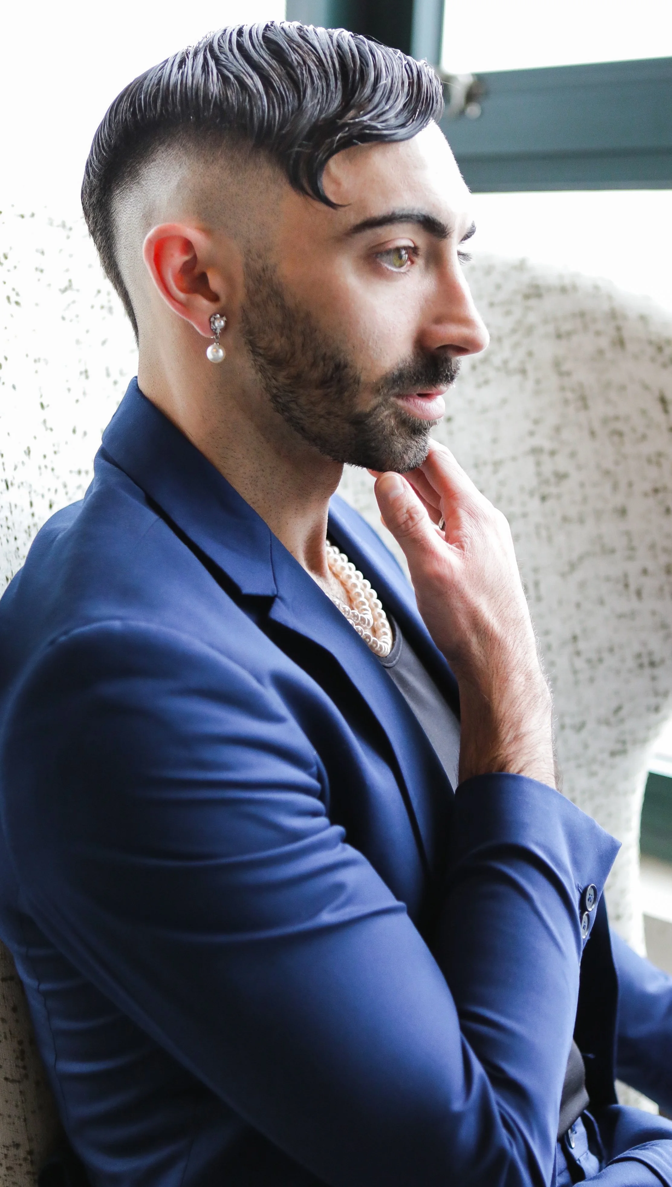 Side profile of a man with styled hair, beard, wearing a pearl necklace, pearl earrings, dark blue blazer, and black shirt, sitting on a light-colored textured chair near a window.