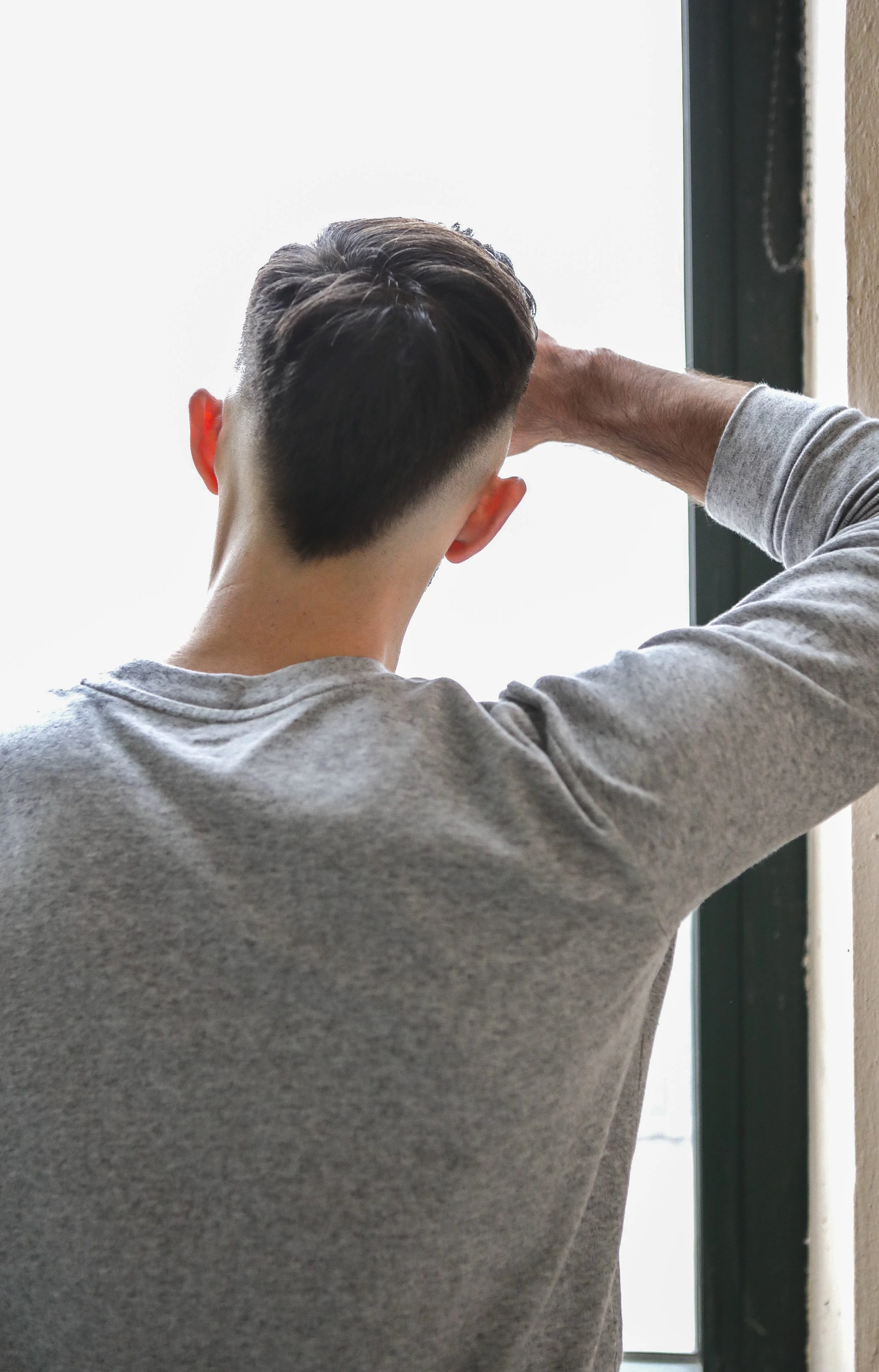 Back of a young man with dark hair, wearing a gray shirt, standing near a window, looking outside with one arm resting on his head.