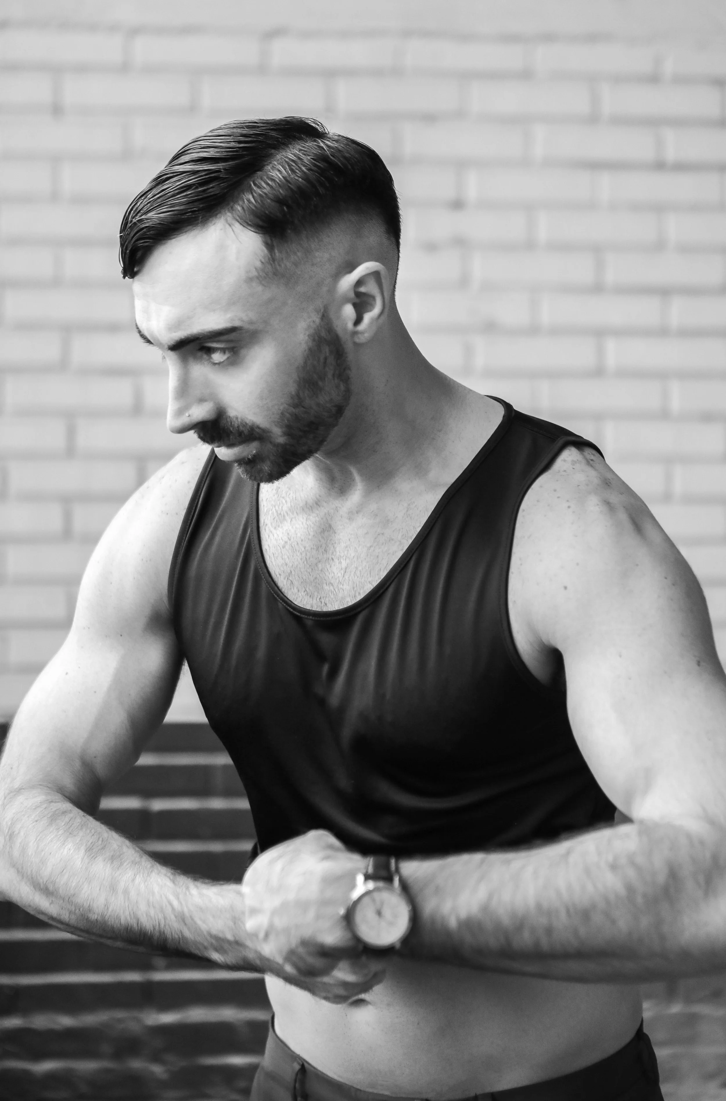 A man with a beard and slicked-back hair, wearing a sleeveless athletic shirt and a watch, checking his wrist in front of a brick wall.