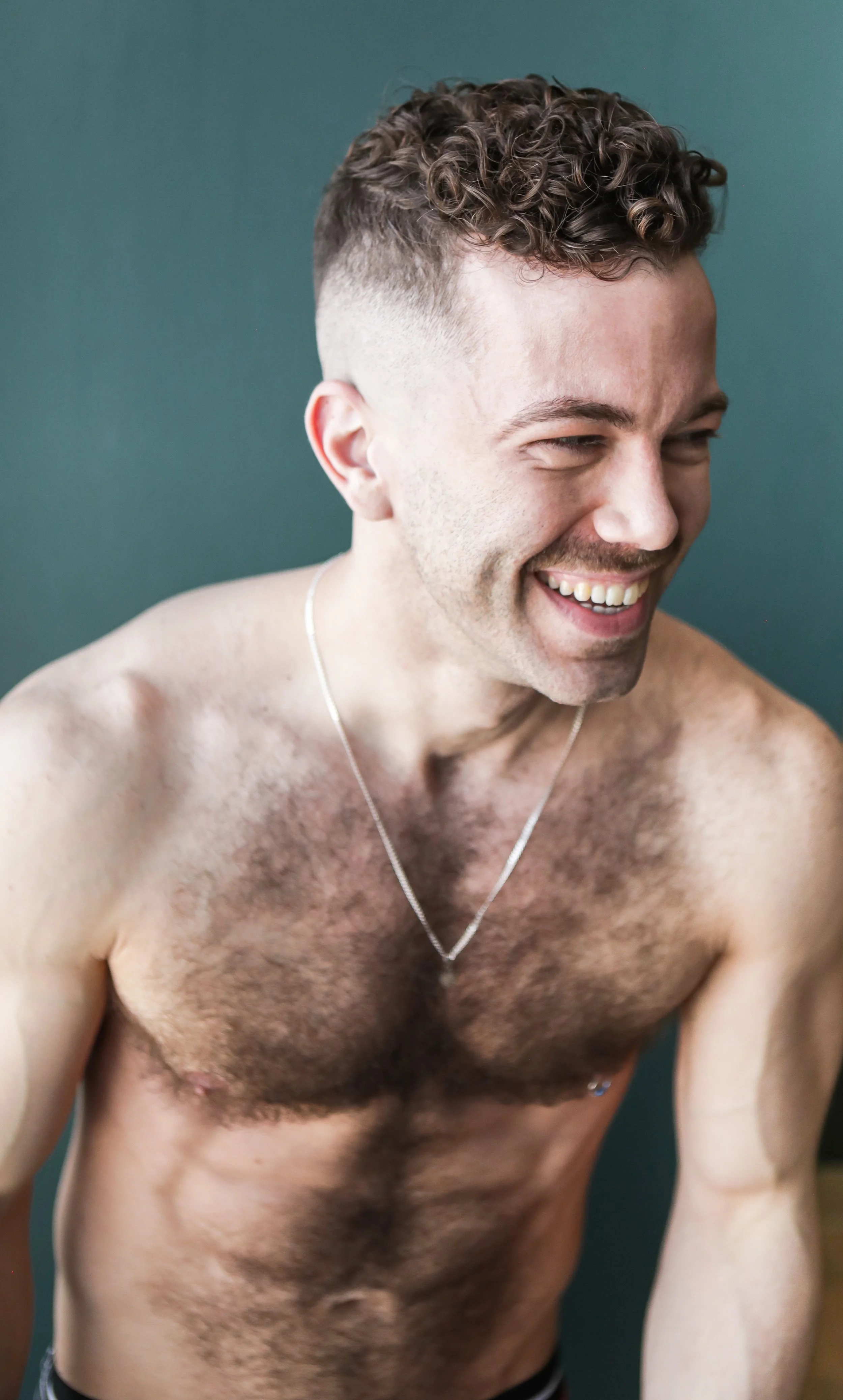 Close-up of a shirtless young man with curly hair, smiling and looking to the side, against a teal background.