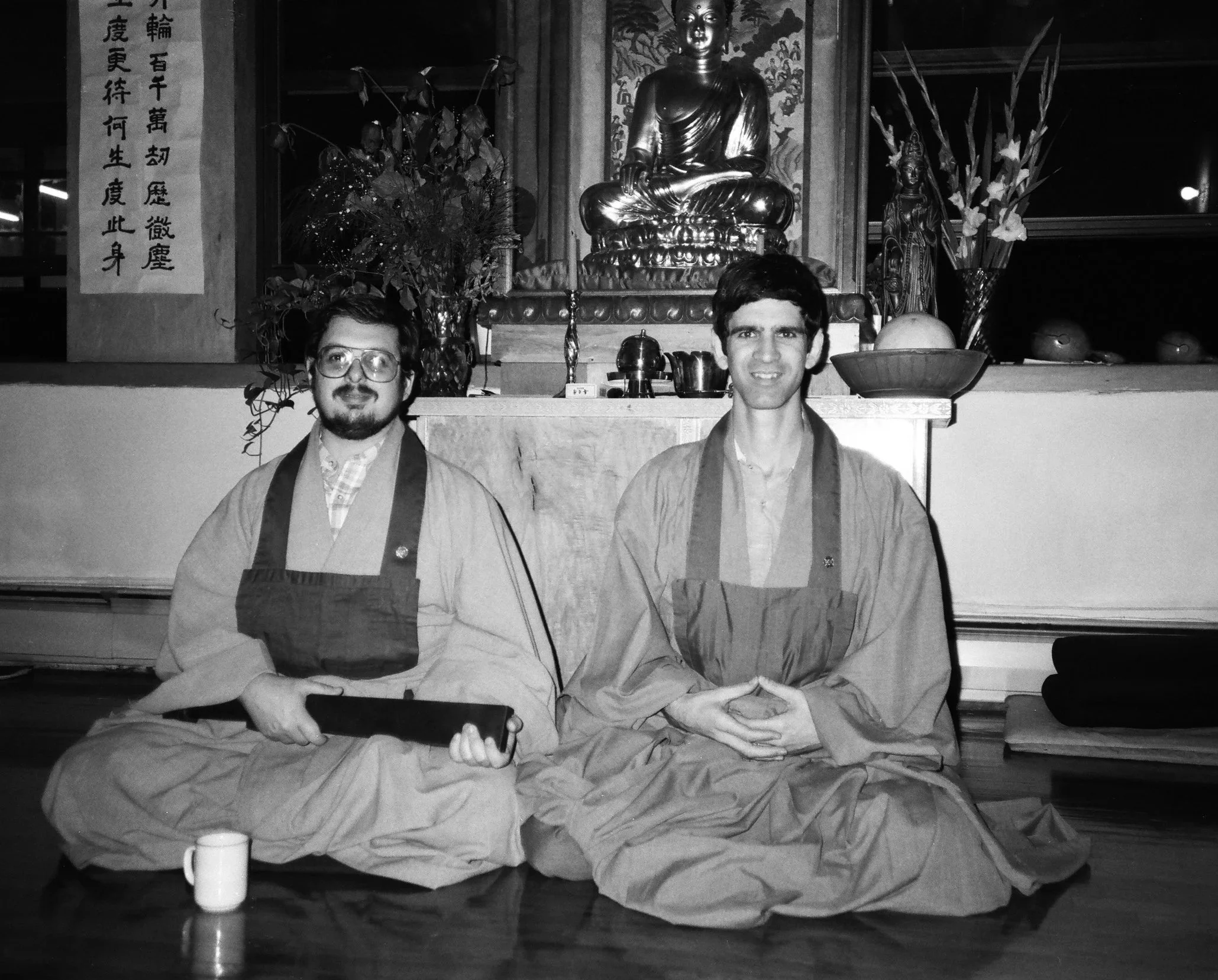 Richard Shrobe and Ken Kessel (left to right) seated in Zen robes in the dharma room at 42 West 17th Street, circa 1985. The Buddha altar and Korean calligraphy are visible behind them.