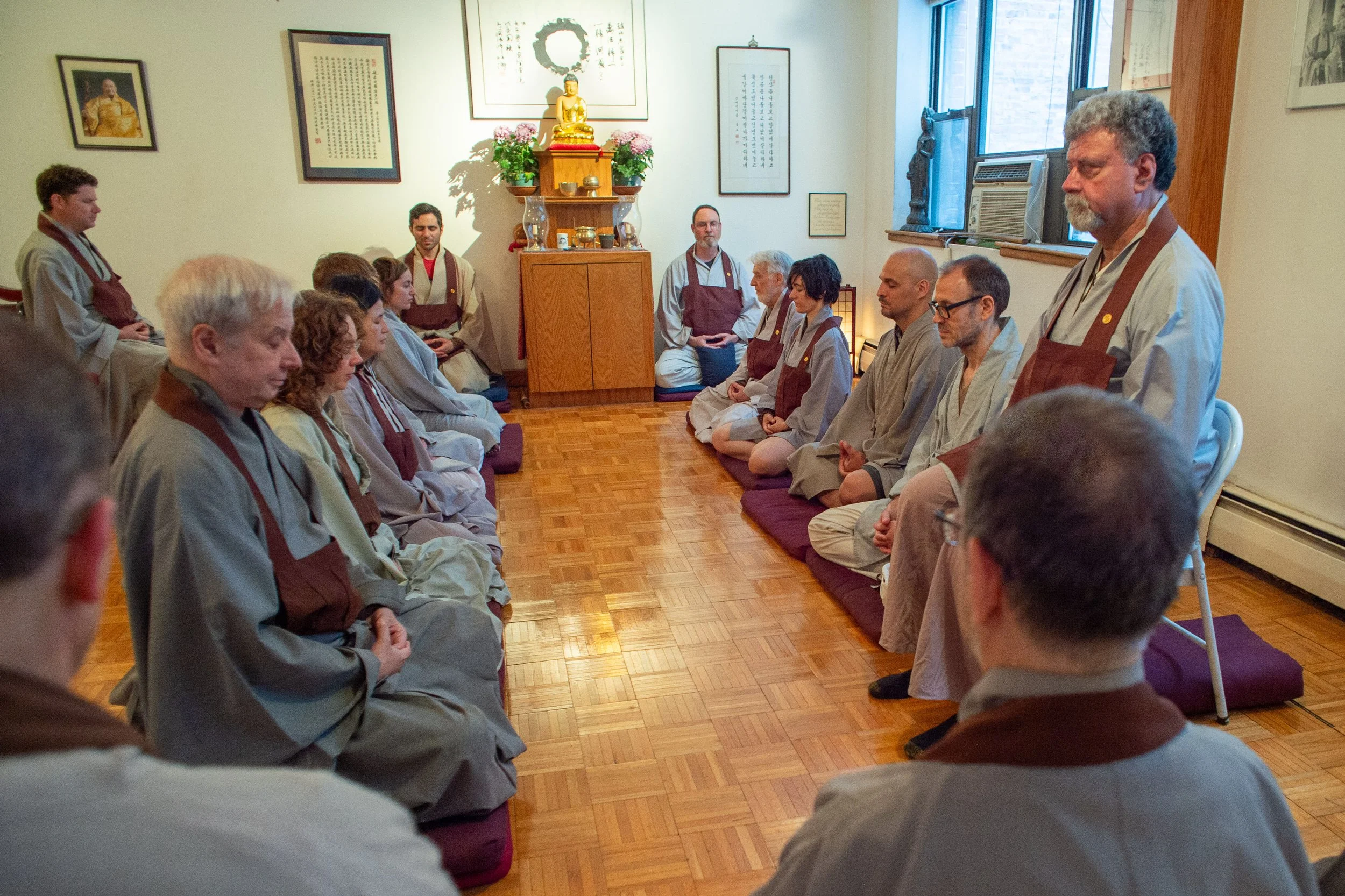 Practitioners in Korean Zen robes seated on rows of mats in the dharma room at 400 East 14th Street, 2016. The Buddha altar with fresh flowers is visible at the front of the room.