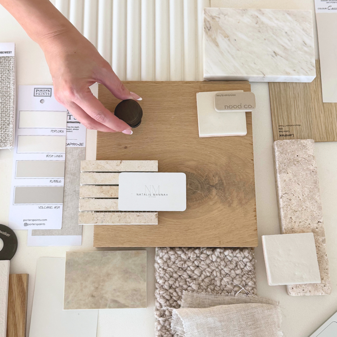 A hand holds a small dark round sample of paint or stain next to various tiles, wood samples, fabric swatches, and color sample cards on a table, likely for interior design or renovation.