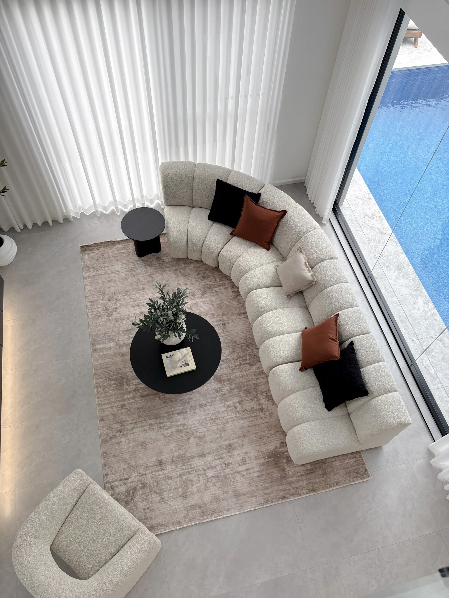 Overhead view of a modern living room with a curved beige sectional sofa, black round side table, light beige armchair, black, brown, and beige throw pillows, a round black coffee table with a plant and a book, and a large window overlooking a blue pool.