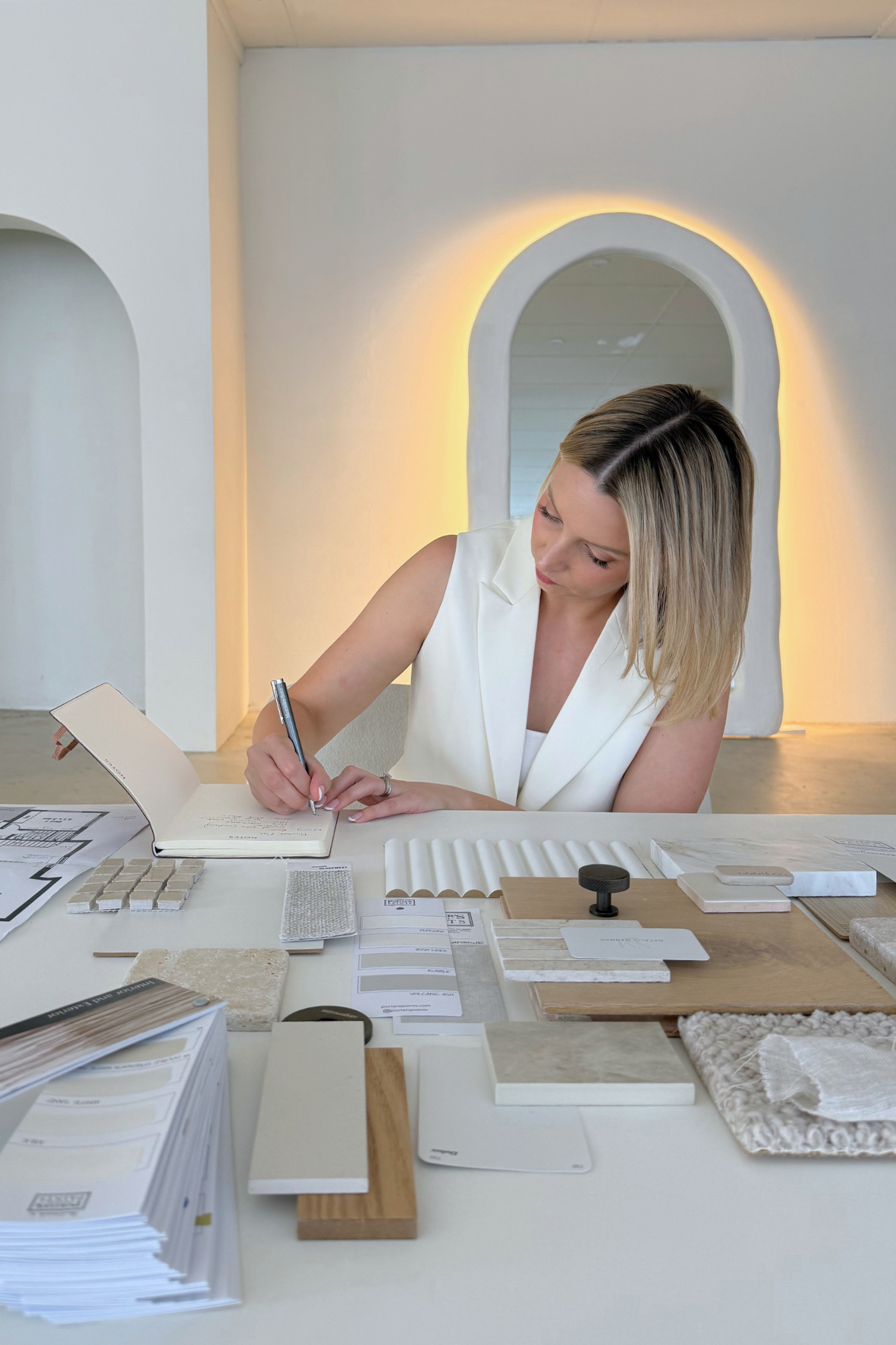 Woman sitting at table with interior design samples, fabric swatches, and color palette swatches, writing in a notebook with modern lighting and architecture in background.