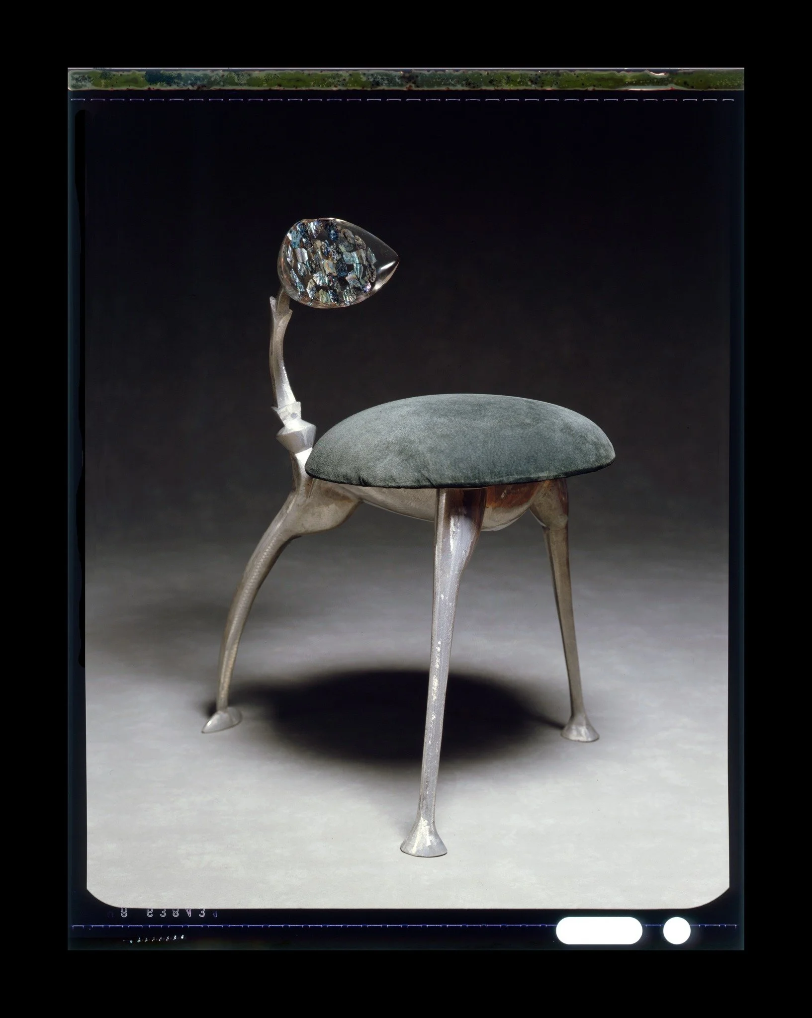 Artistic chair with metallic legs and a green cushioned seat, featuring an ornate backrest with mosaic detailing on a dark background.