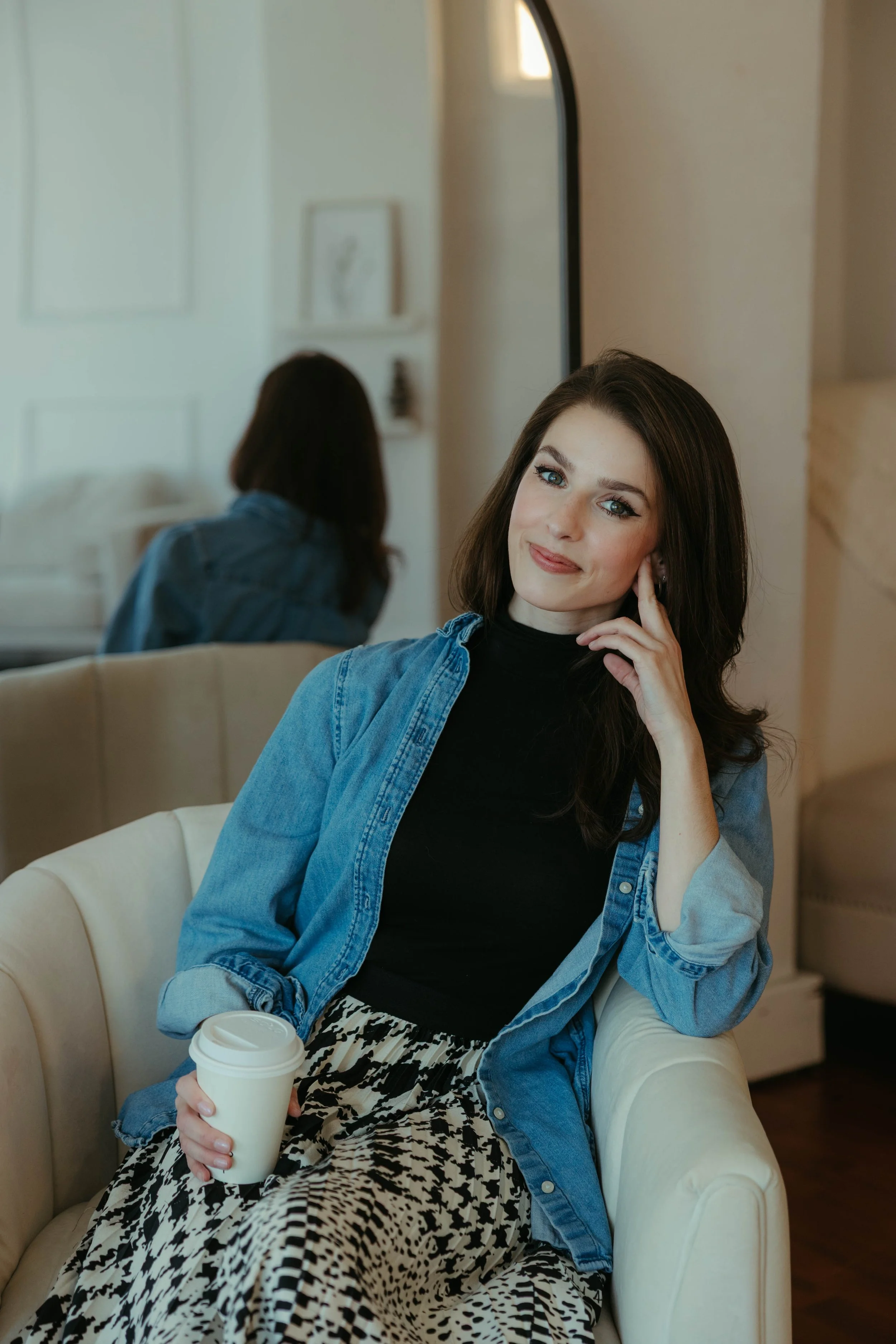A woman sitting on a beige couch, holding a white takeout coffee cup, smiling, with dark brown hair, wearing a black turtleneck and a blue denim shirt, in a cozy room with a mirror and artwork in the background.