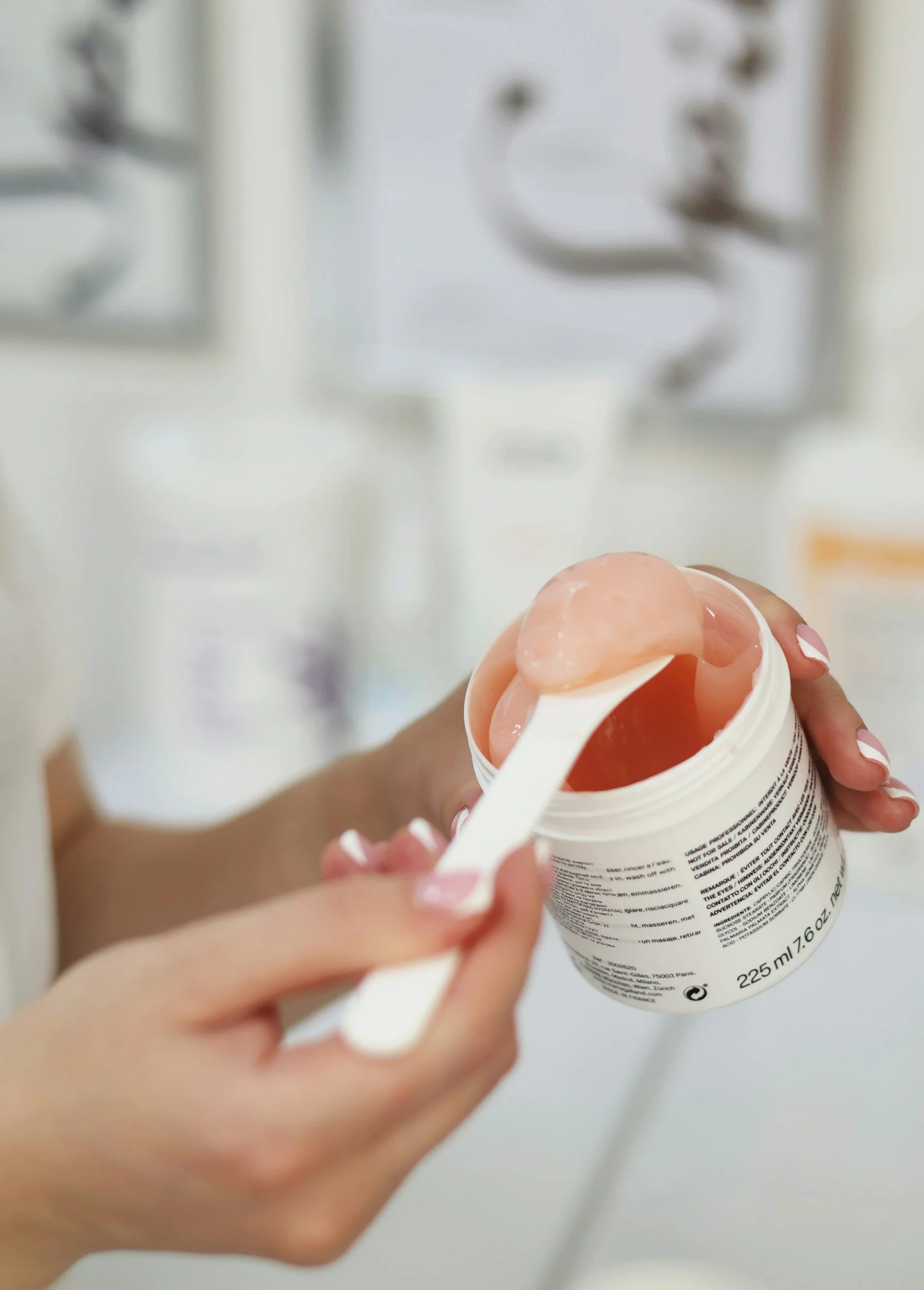 Person holding a white container with pink gel inside, using a white spatula to scoop out the gel. In the background, blurred, are wall decorations.