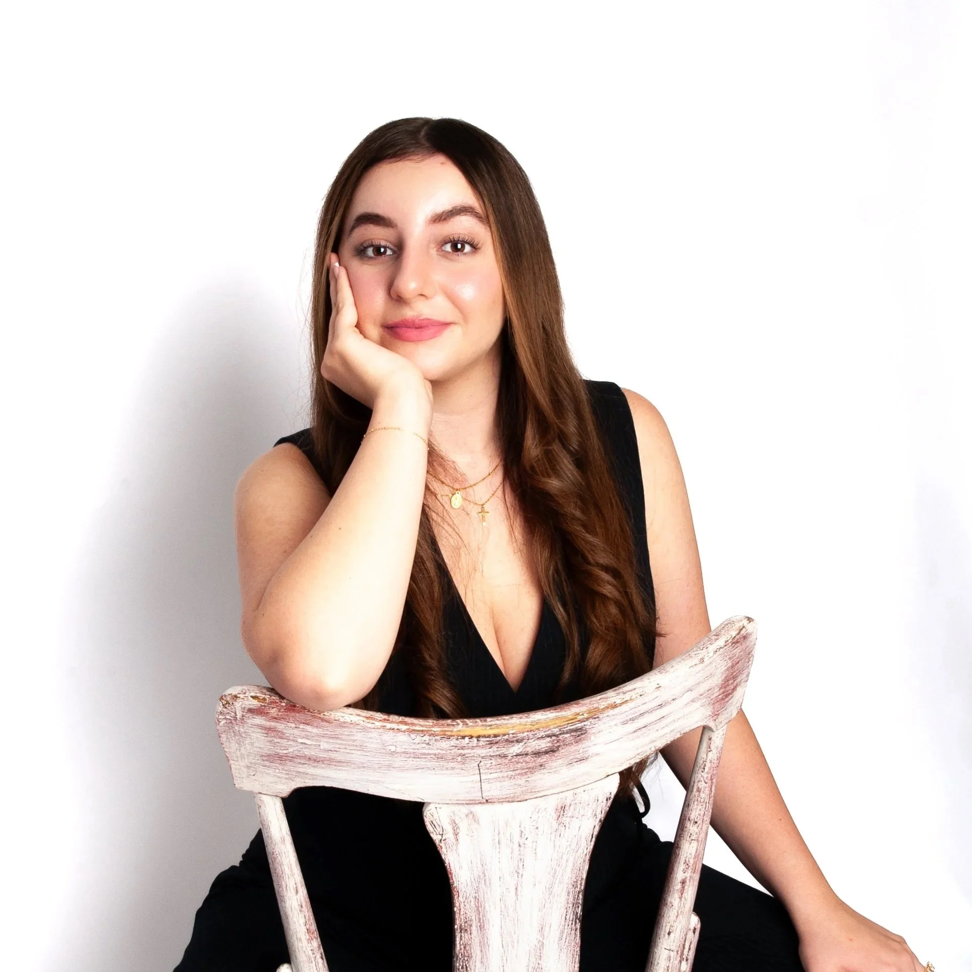 A young woman with long brown hair sits behind a rustic white wooden chair, resting her face on her hand, looking at the camera, against a plain white background.