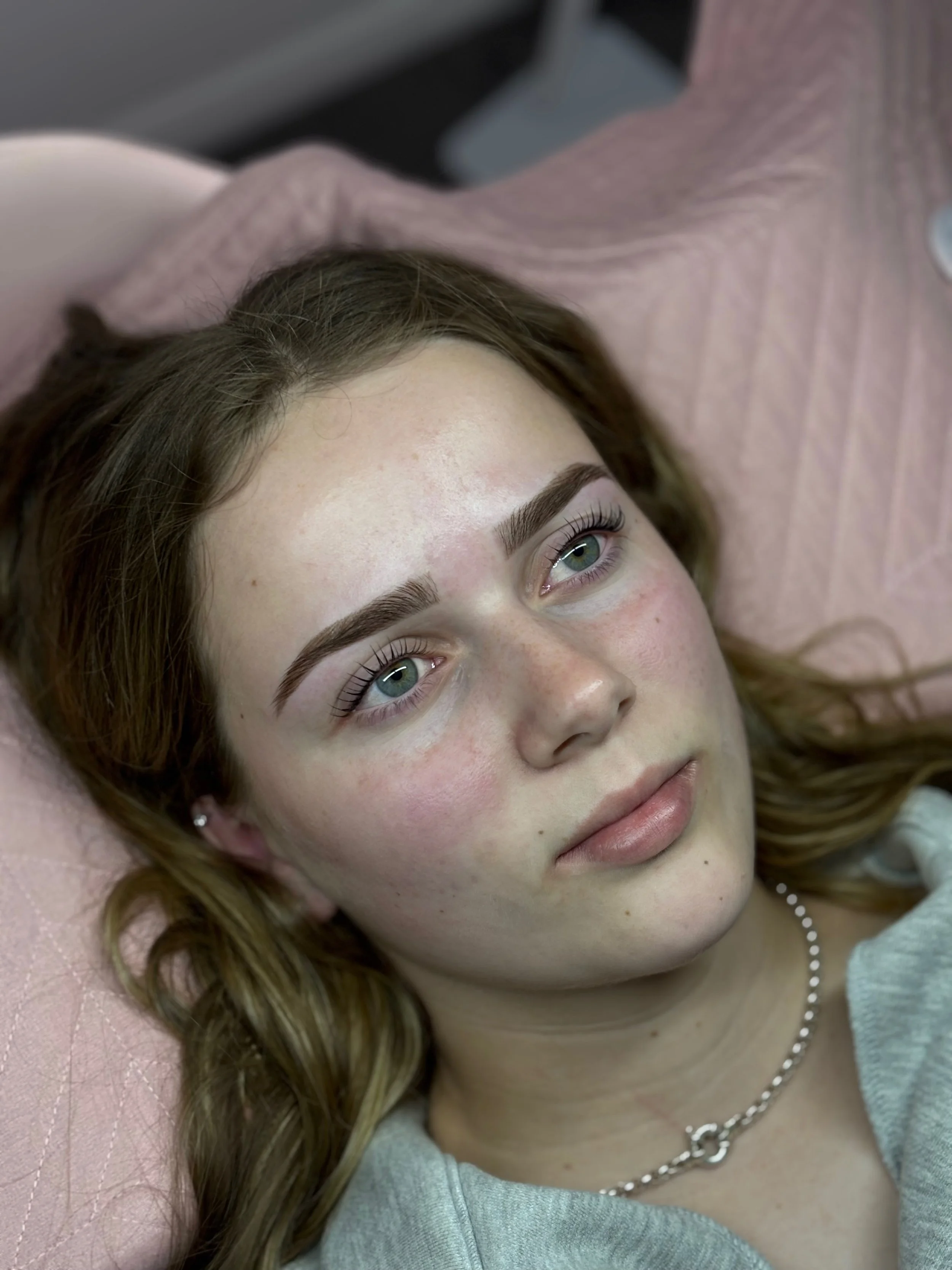 Close-up of a young woman lying down, looking up, with brown hair, blue eyes, and a silver necklace, in a relaxed setting.