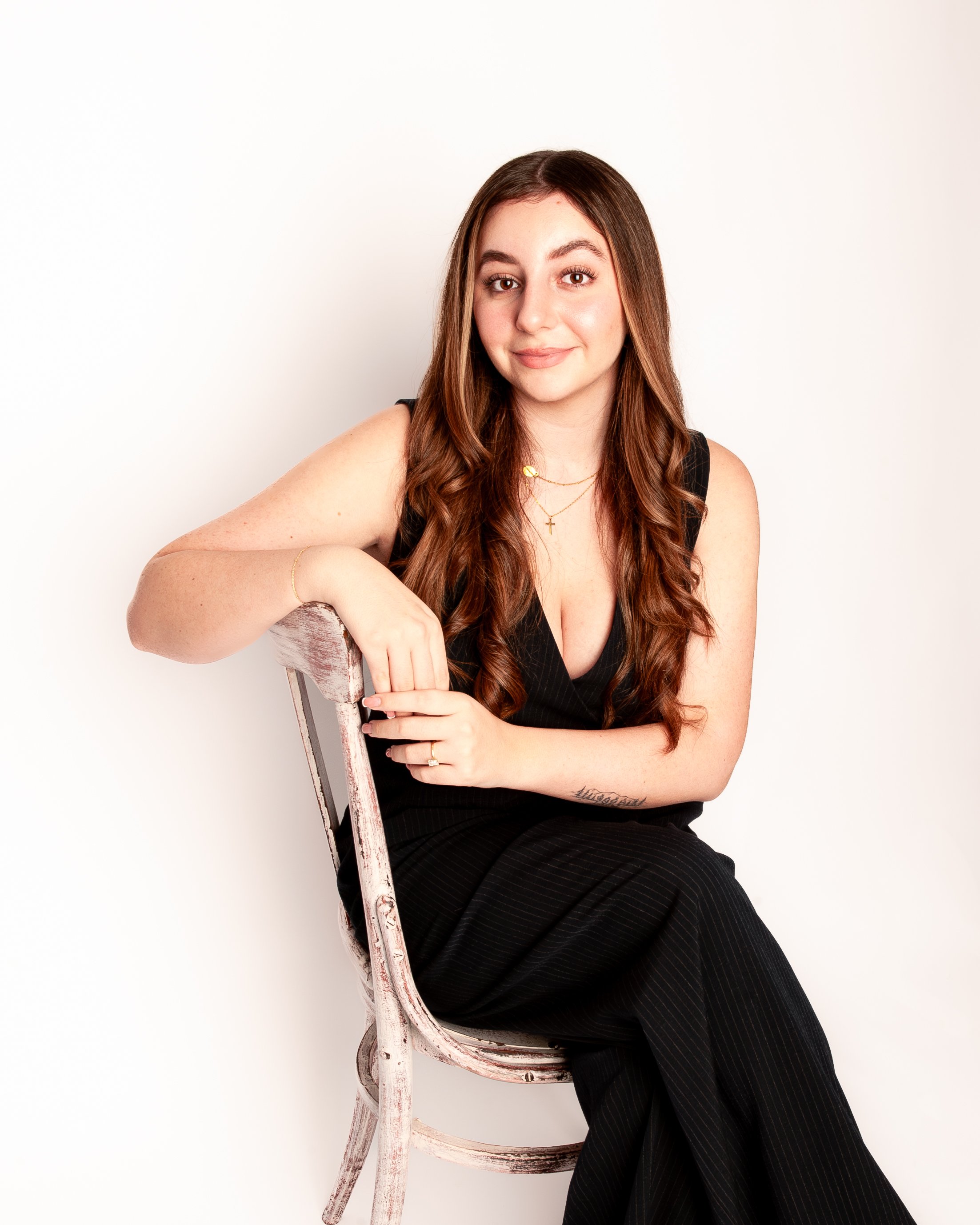 Young woman with long, wavy brown hair, wearing a black tank top and layered necklaces, sitting on a rustic wooden chair against a white background.