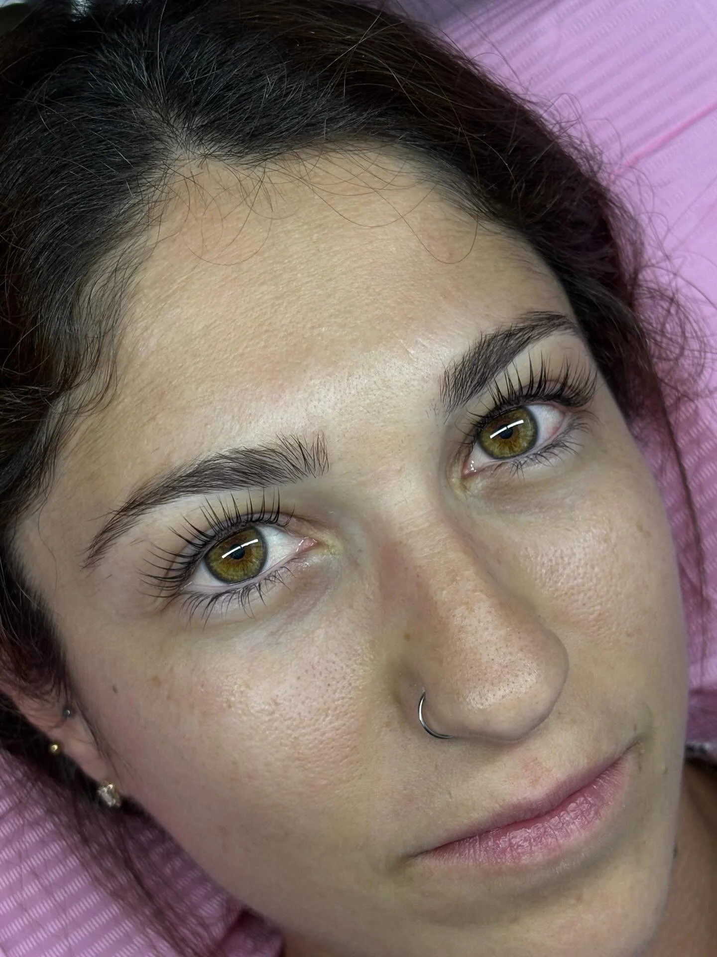 Close-up of a woman's face showing hazel eyes, well-groomed eyebrows, and light skin with freckles, lying on pink fabric.