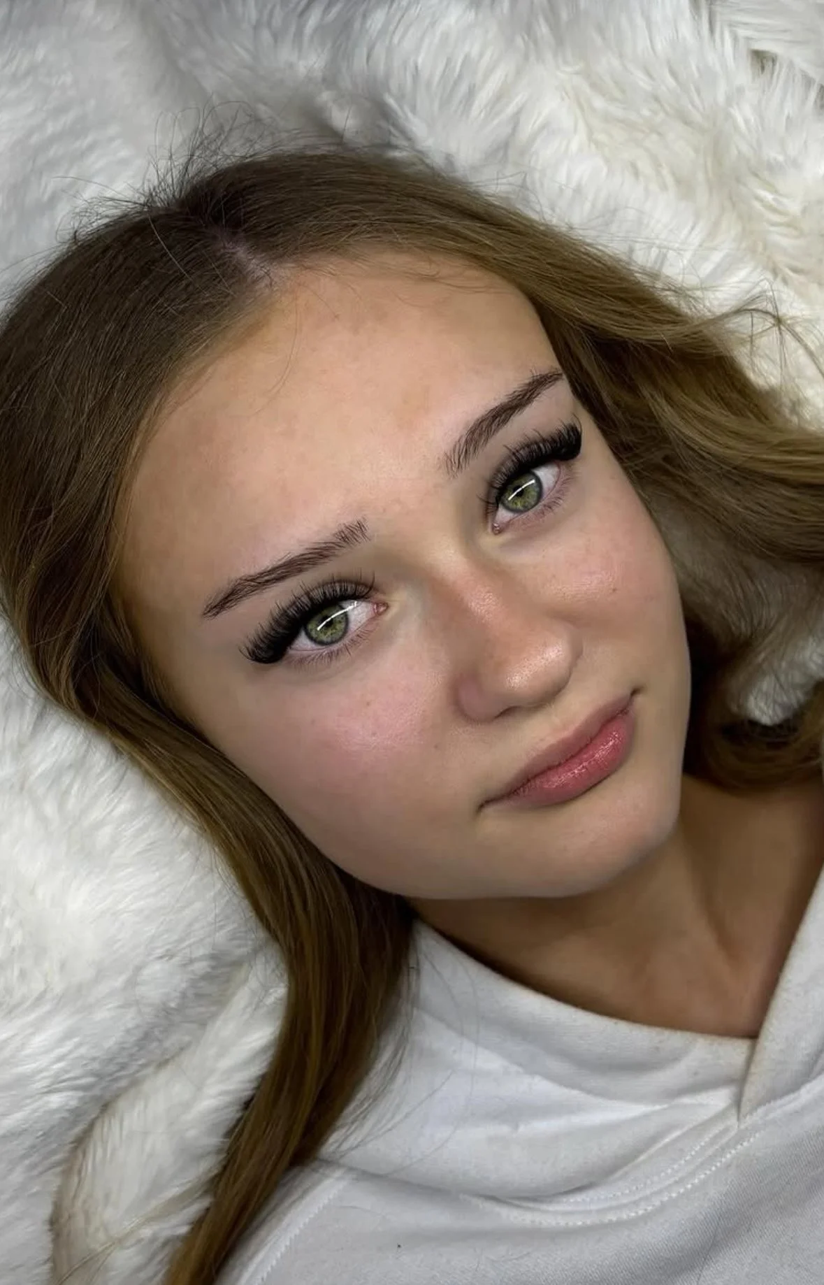 Close-up of a young woman with green eyes, long eyelashes, with her head resting on a white fluffy surface, wearing a white top.