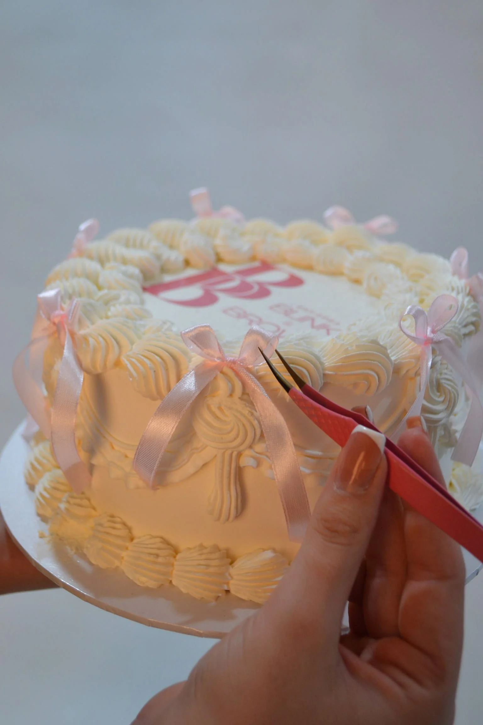 A person holding a cake decorated with white icing and pink ribbons, using a small knife to cut a slice.