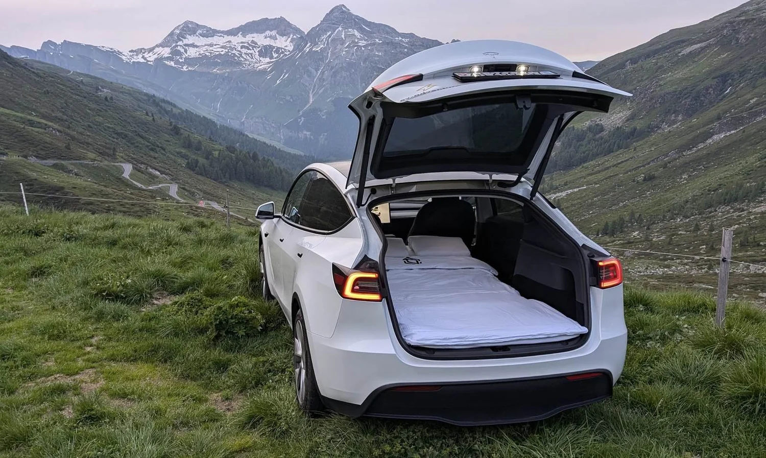 Weisses Tesla Model Y mit offener Heckklappe und Matratze im Kofferraum vor einer Bergkulisse