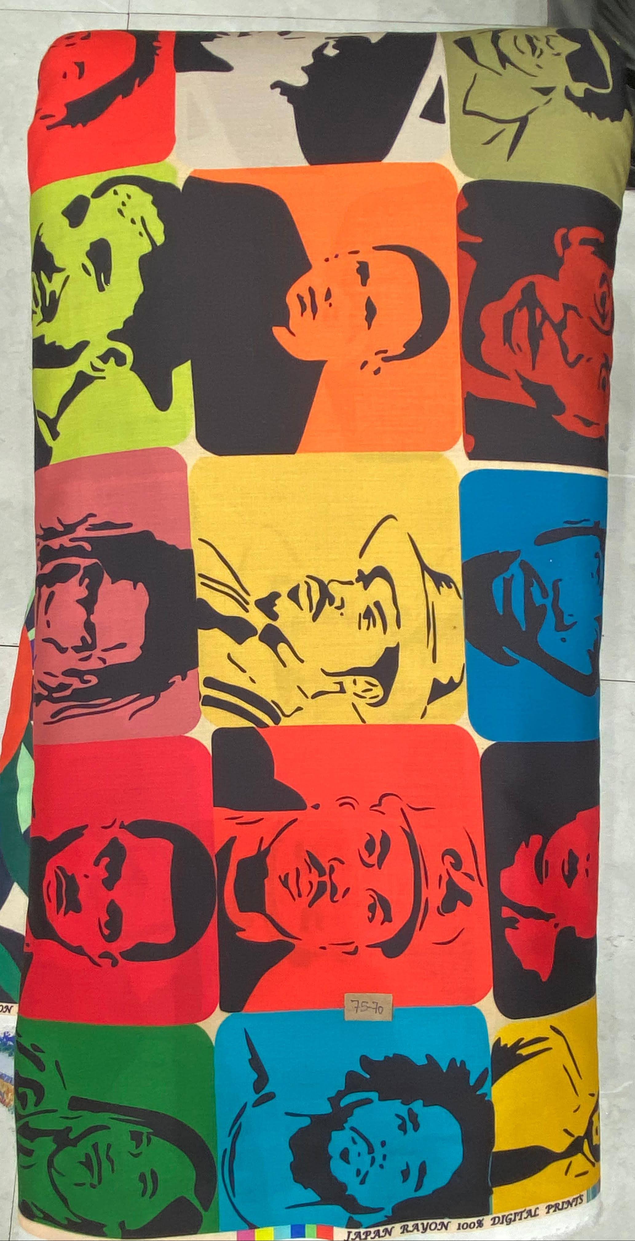 Vibrant multi-colored fabric featuring an expressive pop-art style design with stylized faces, bold geometric shapes, and rich golden-yellow, blue, and black tones.