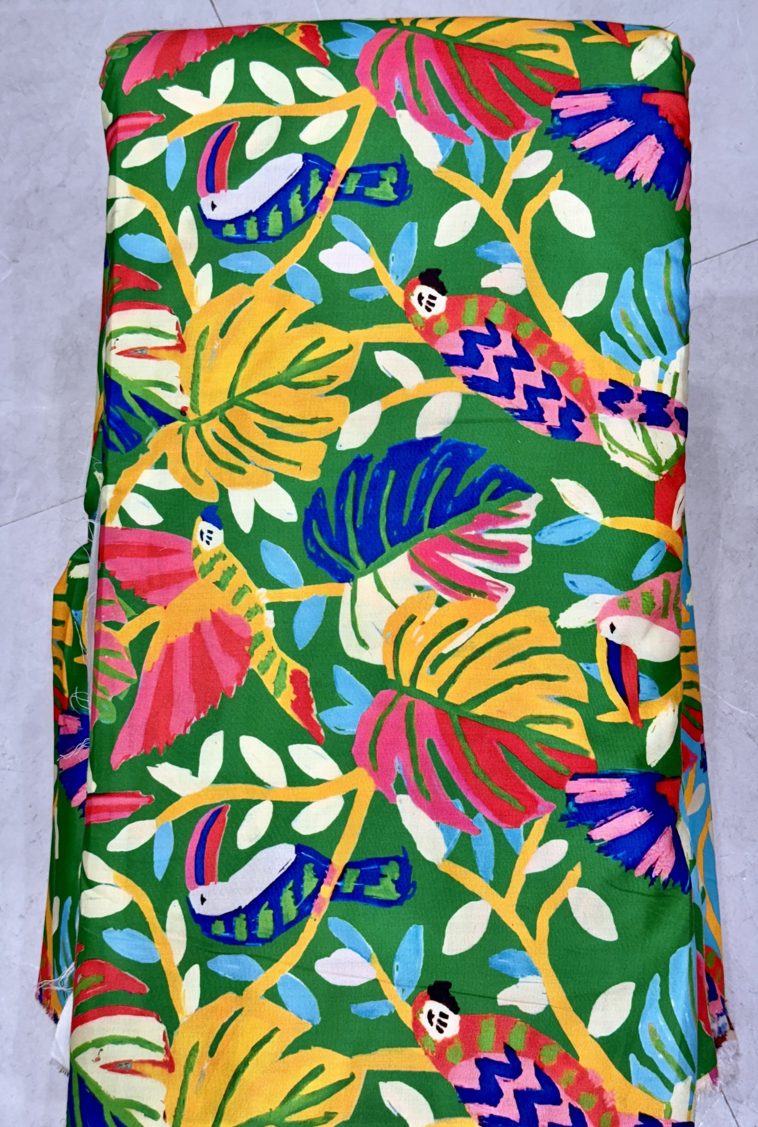 Vibrant green fabric featuring a lush tropical print with colorful exotic birds, vibrant floral blossoms, and detailed botanical palm leaves.
