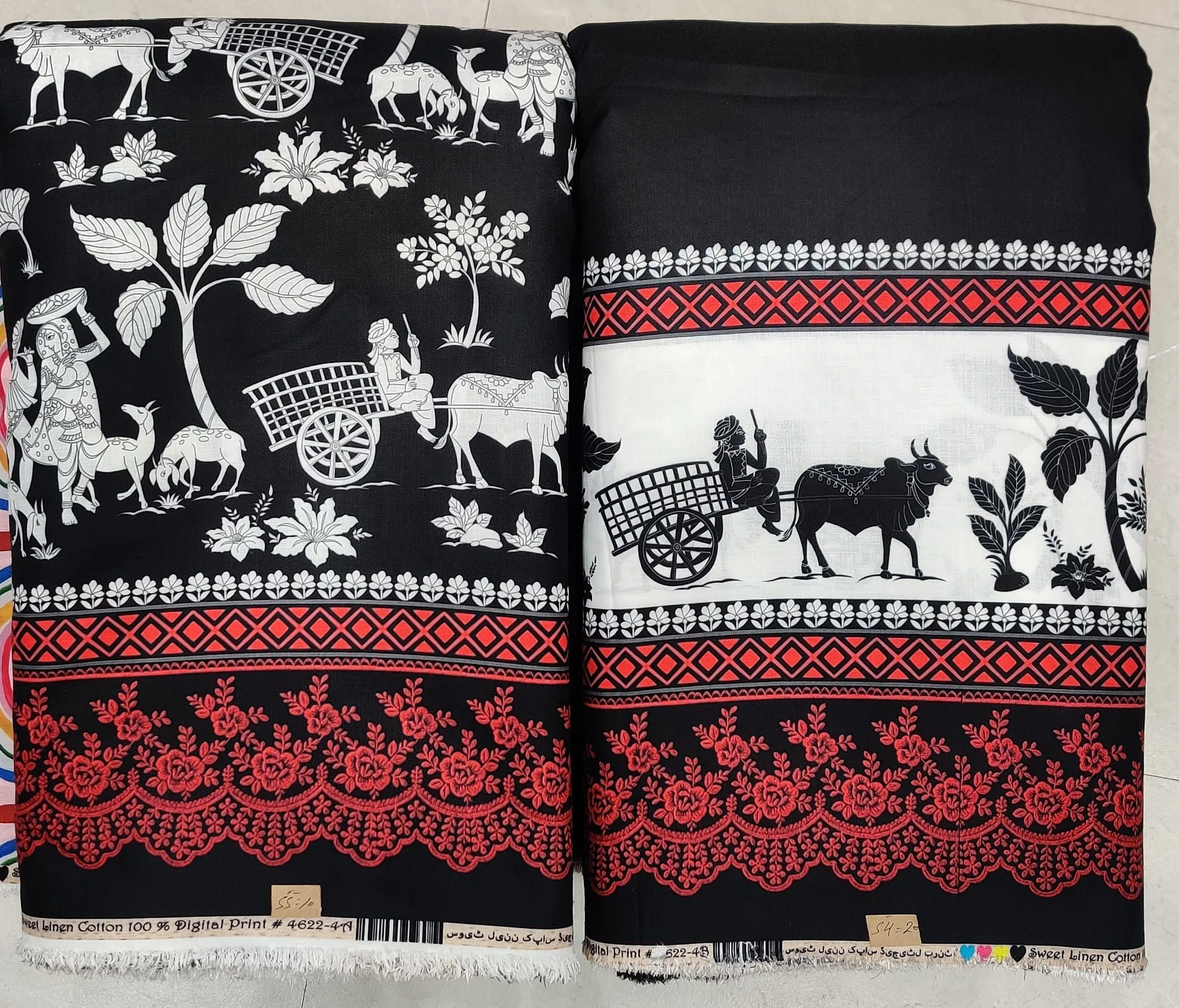 Rich jet-black and pristine ivory cotton fabric featuring a traditional folk-art village scene with human figures, ox carts, and palm trees along the main panel, finished with a detailed geometric and vibrant red embroidered-style border.