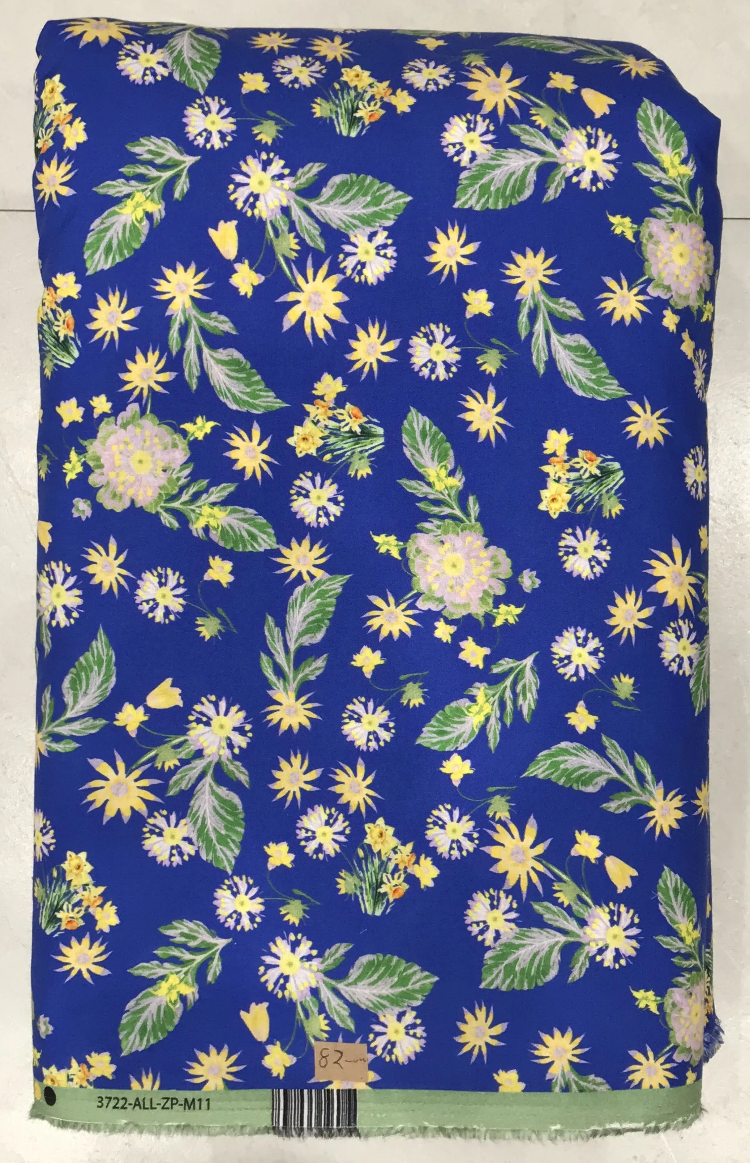 Rich royal blue background featuring a dense, vibrant floral print with delicate yellow, white, and soft blue blossoms interwoven with lush green leaves.