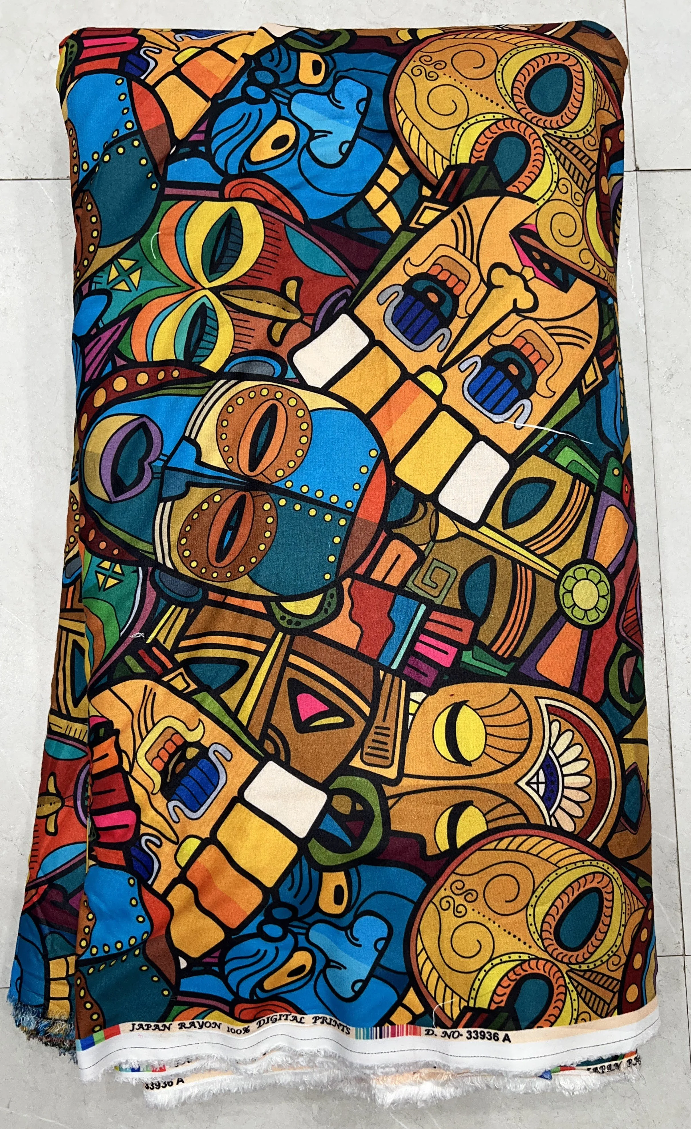 Rich multi-colored fabric featuring an intricate tribal mask pattern with bold geometric motifs, tribal artwork elements, and vibrant shades of blue, orange, and gold.