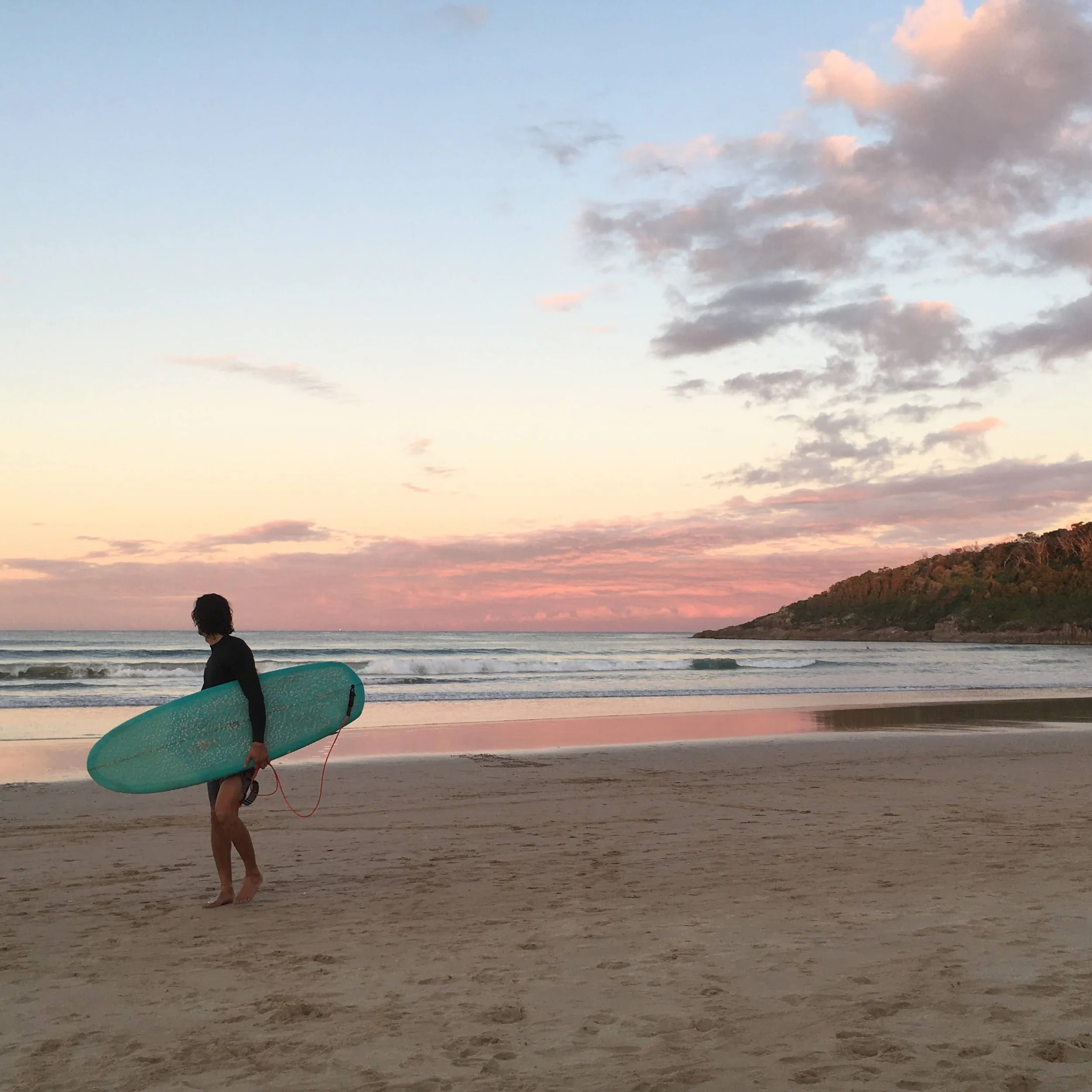 Une personne avec une planche de surf marchant sur la plage au coucher du soleil, avec le ciel nuageux et la mer calme en arrière-plan.