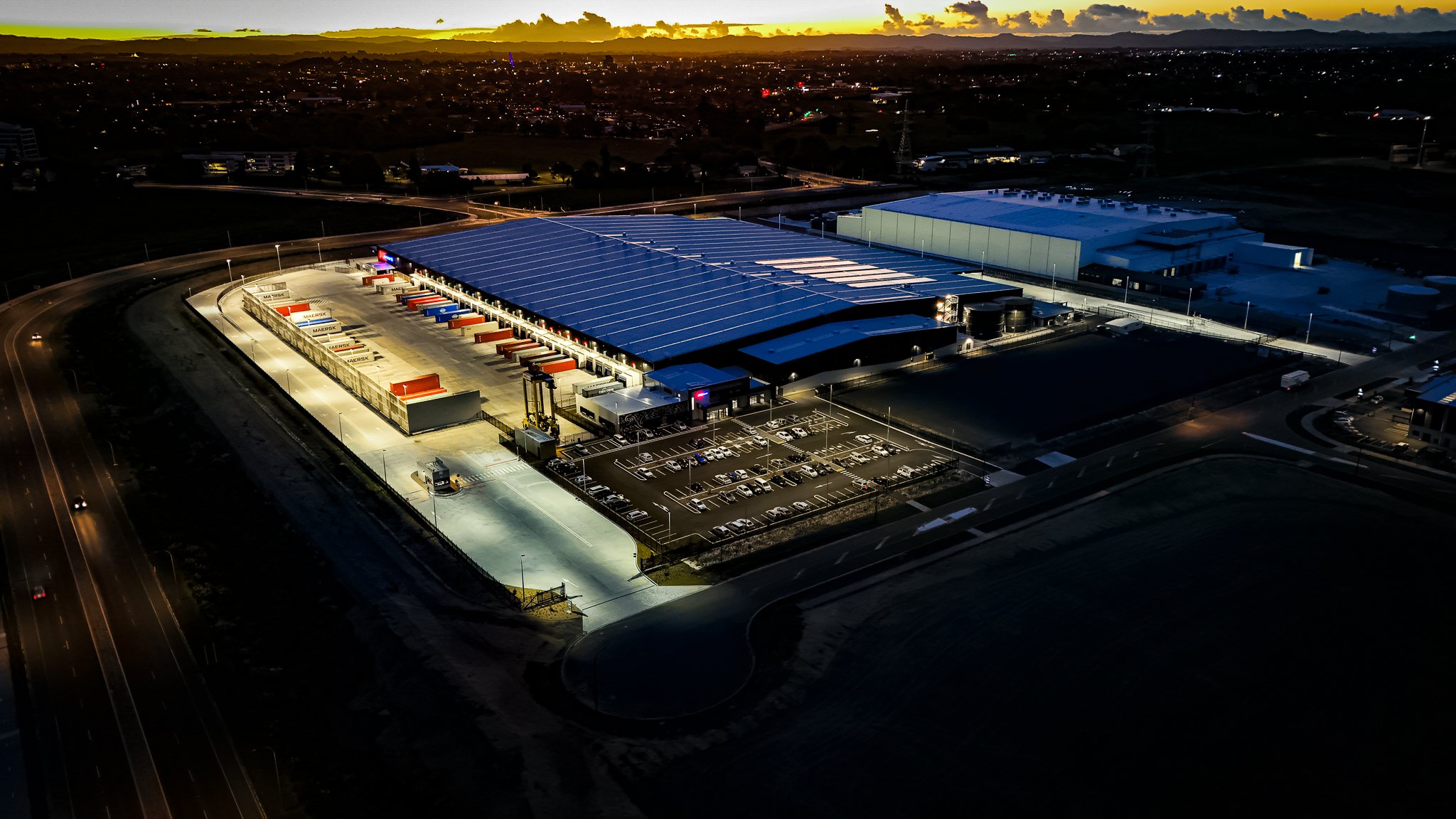 An aerial view at dusk of a large warehouse or distribution center with trucks parked at loading docks, a parking lot with cars, and surrounding roads and fields.