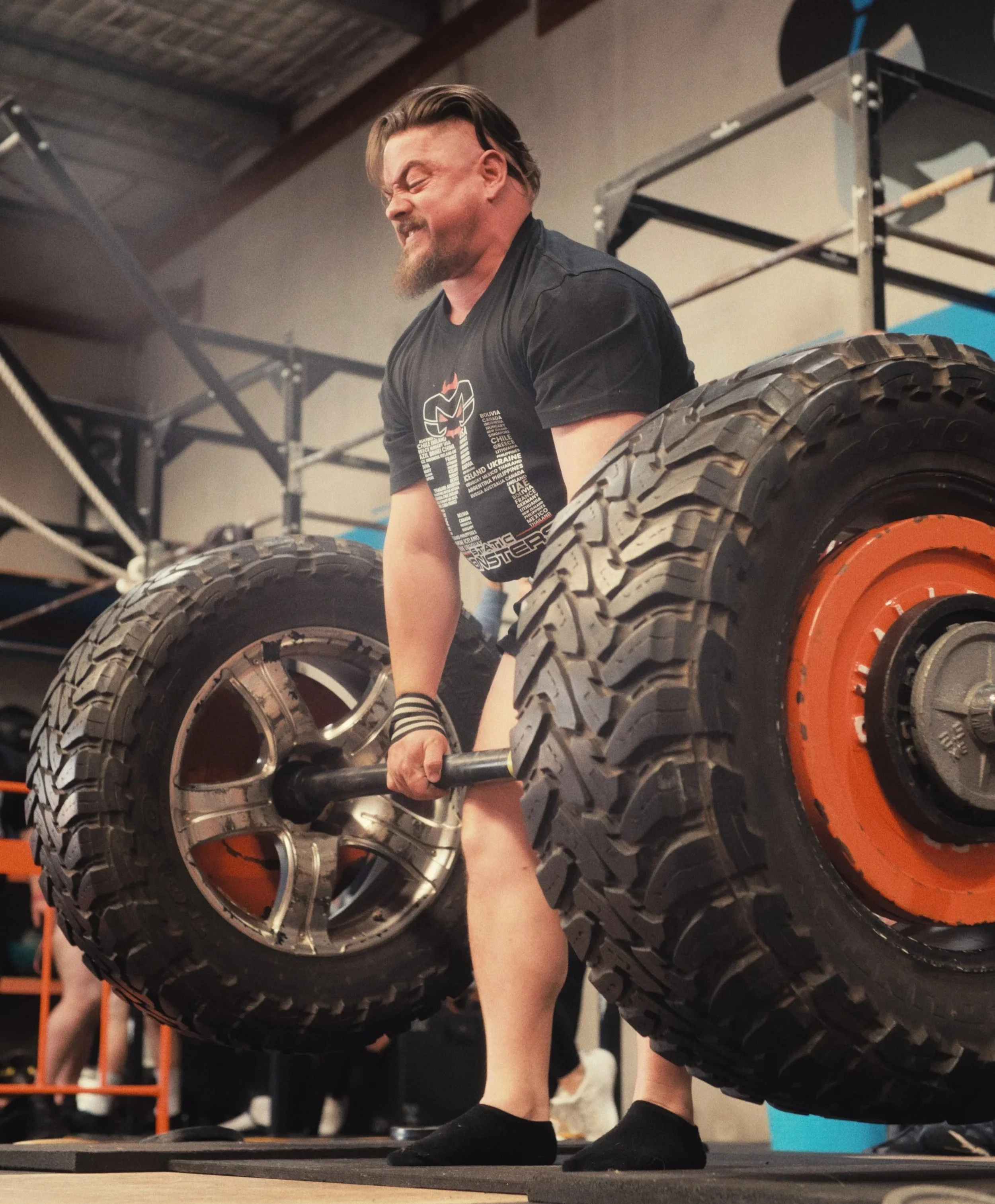 A man with a beard and long hair, wearing a black T-shirt, is lifting a large tire with a weightlifting bar in a gym setting. The background includes gym equipment and a high ceiling.