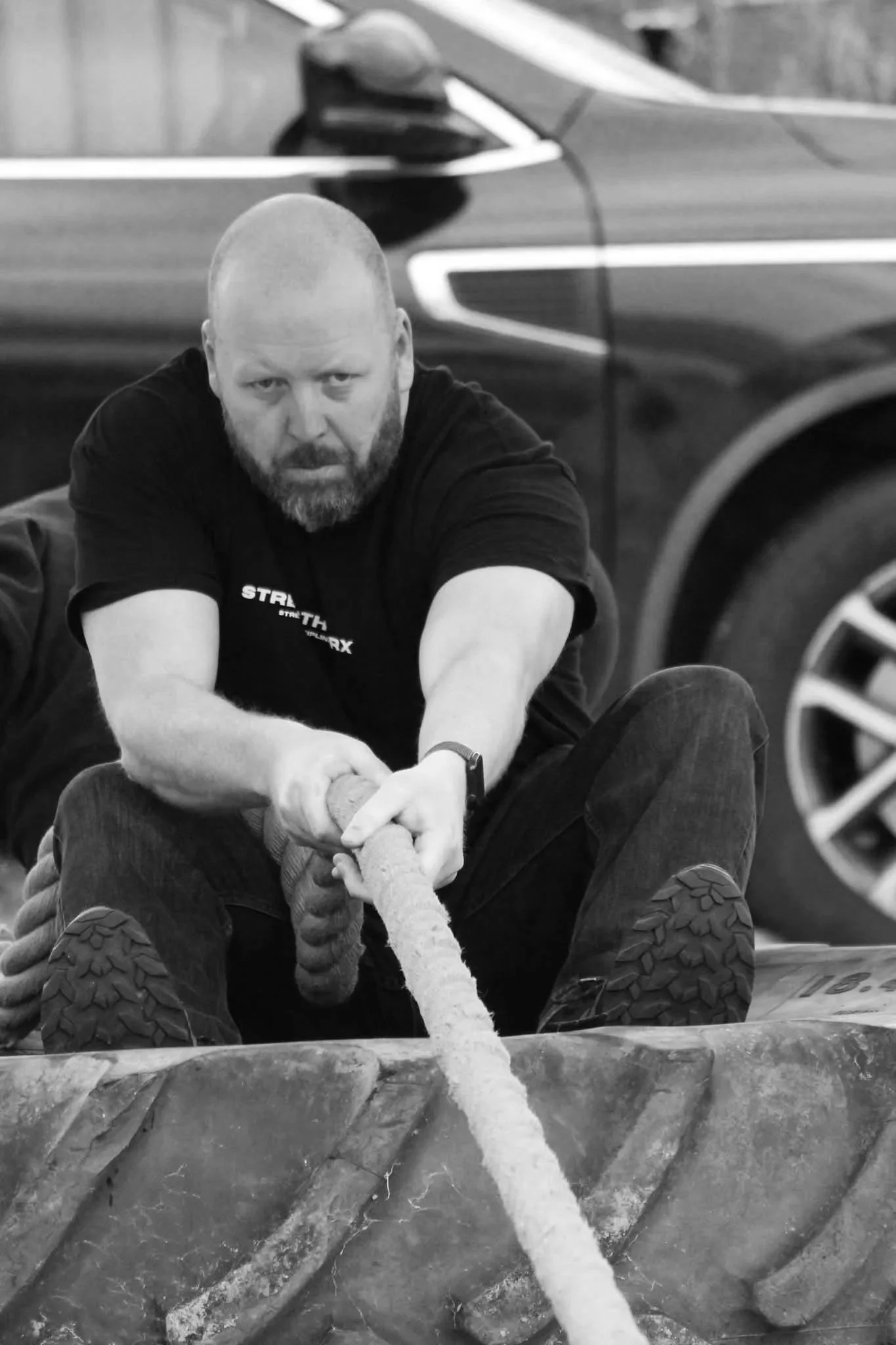 A man with a beard and shaved head pulling a thick rope during a workout or fitness activity, with a car in the background.