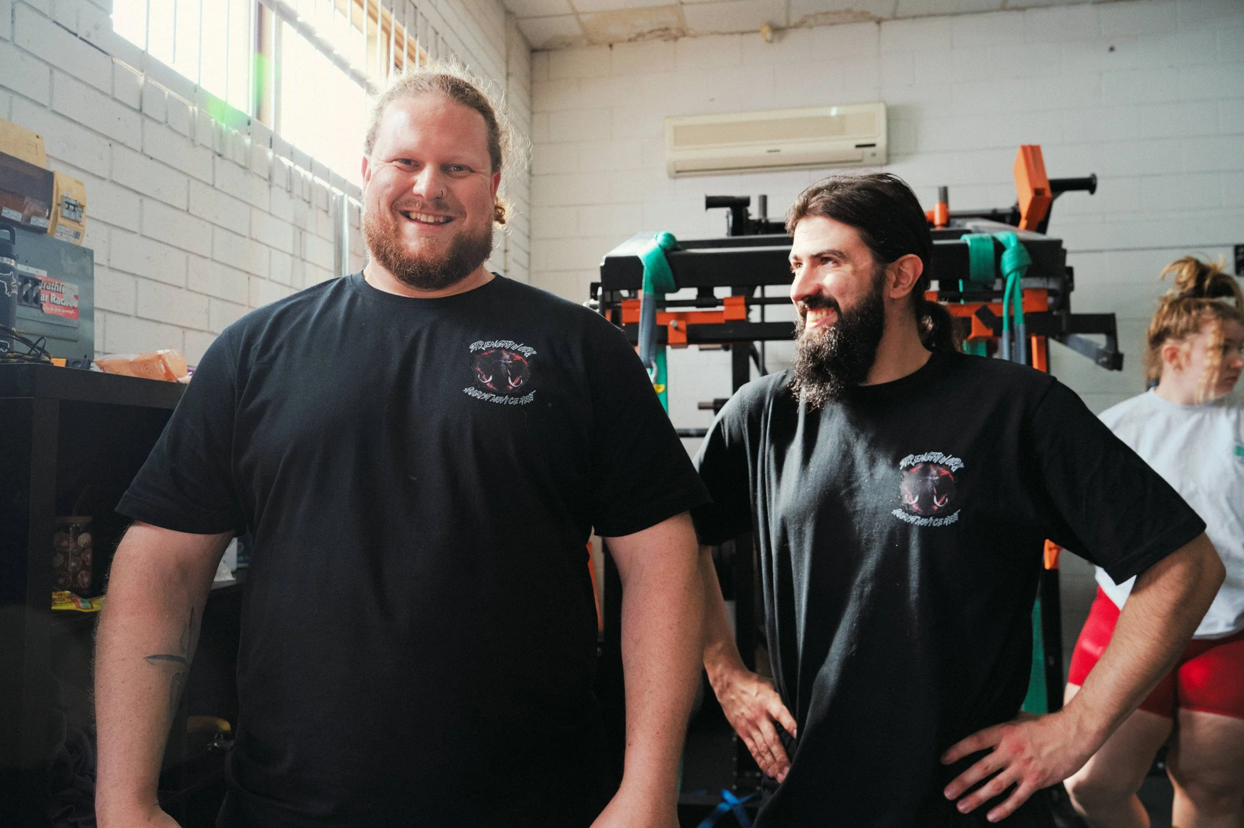 Two men standing inside a gym or training facility, smiling and talking to each other, with fitness equipment in the background.