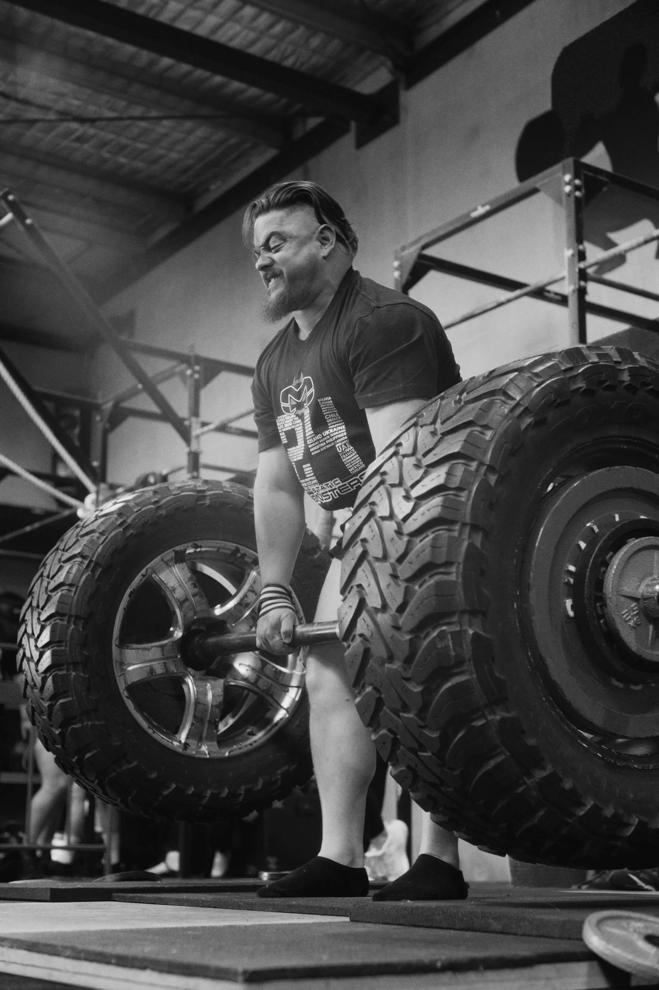 A man lifting a large tire in a gym, smiling, with weightlifting equipment and other gym members in the background, in black and white.
