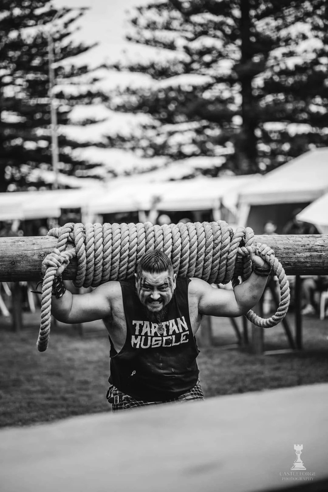 A muscular man with face paint, wearing a tank top with the words 'Tartarian Muscle,' is holding a large wooden log with thick rope wrapped around it on his shoulders, participating in an outdoor strength competition.