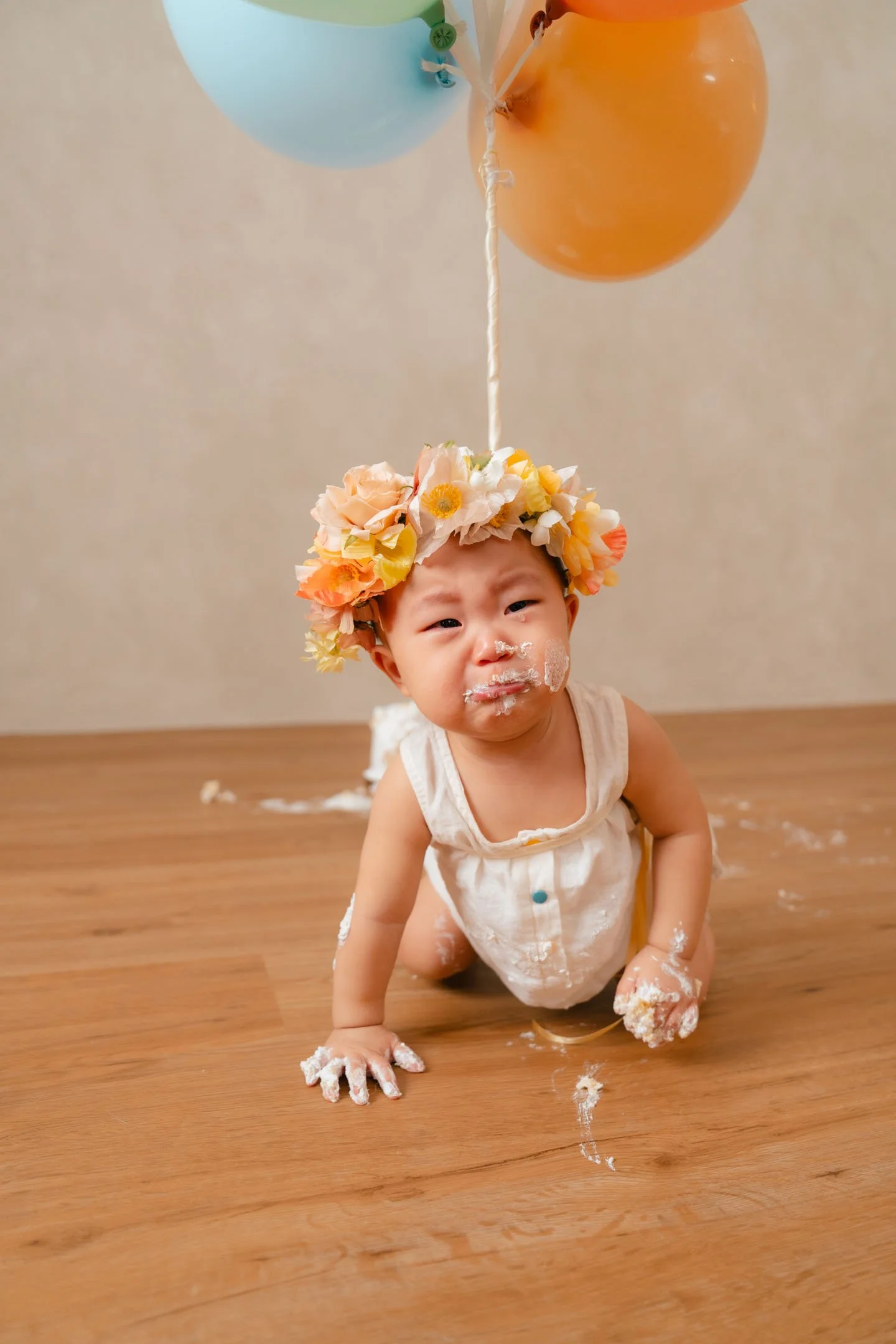 Young child wearing a flower crown, crawling on wooden floor with cake and frosting on face and hands, under colorful balloons, during a birthday celebration.