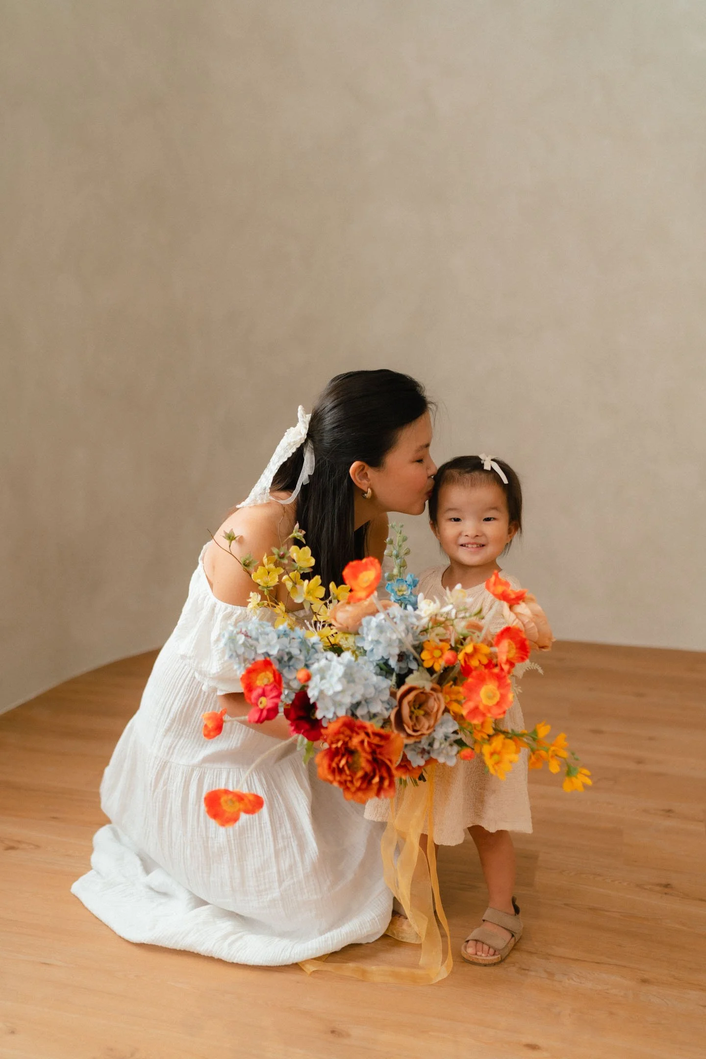 A woman in a white dress kneels and kisses a young girl in a beige dress holding a colorful bouquet of flowers. The girl is smiling, and both have hair adorned with white ribbons, standing on a wooden floor against a plain beige wall.
