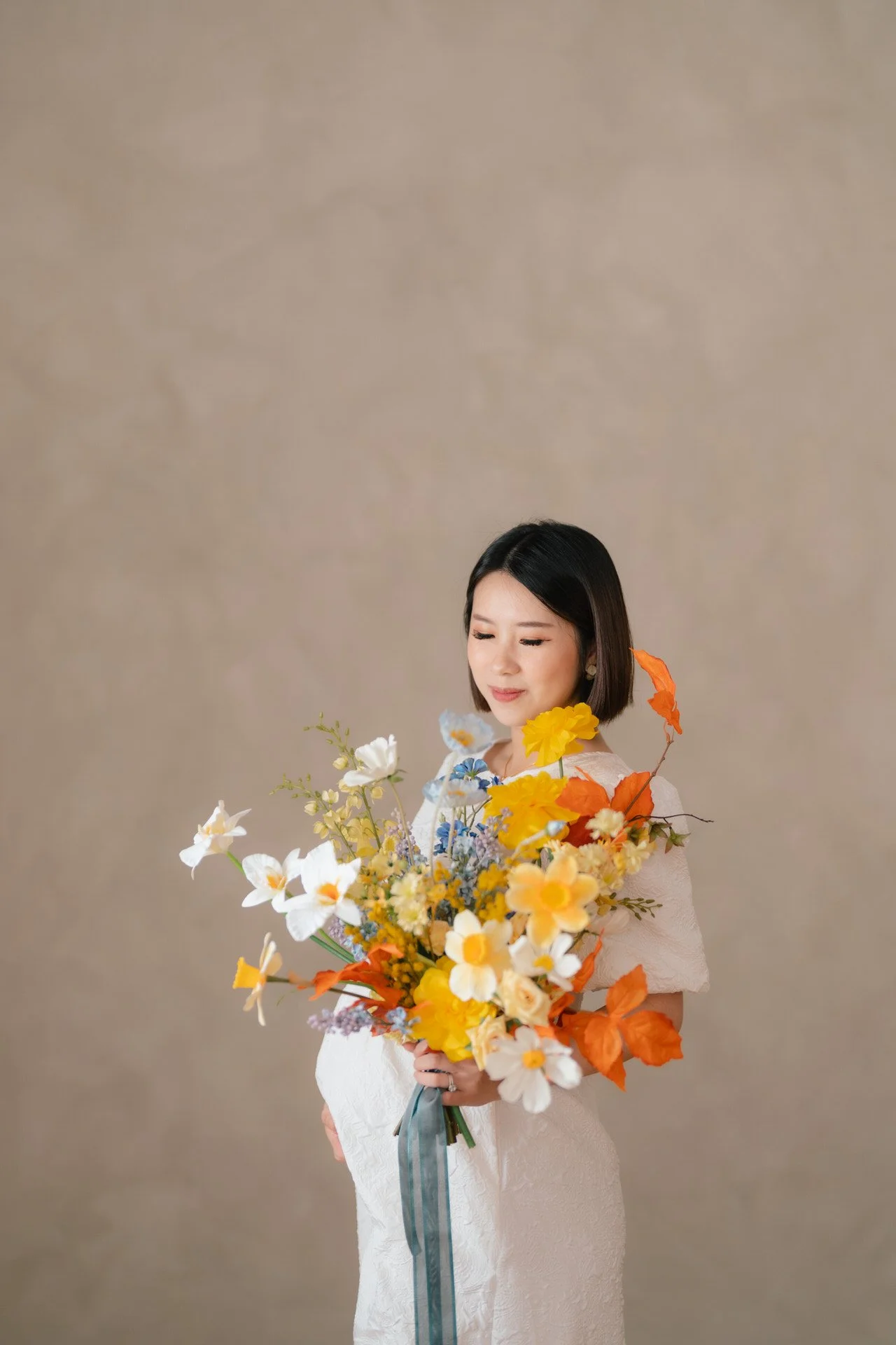 A woman with short black hair holding a large bouquet of various colorful autumn flowers and leaves, standing against a plain, light brown background.