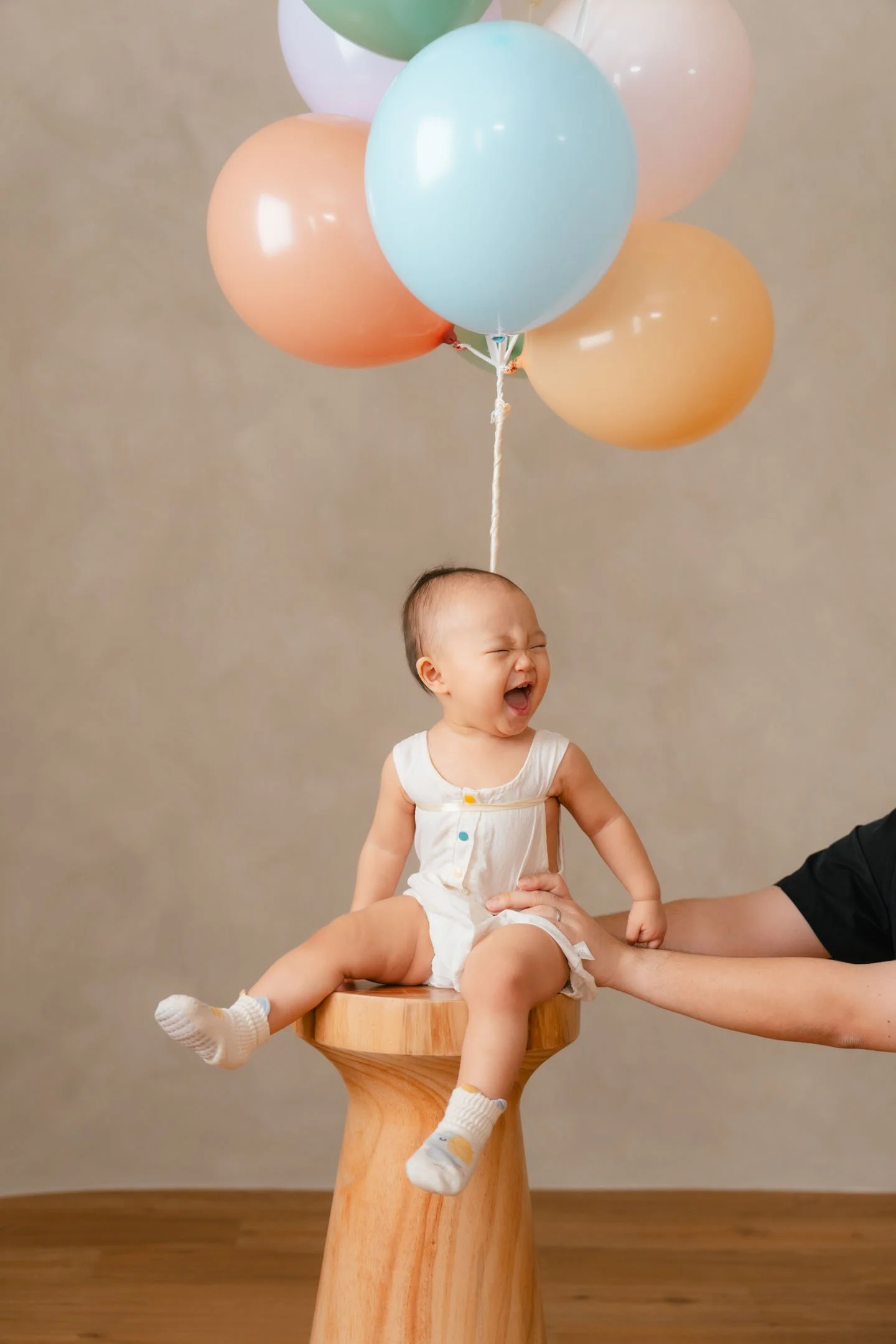 A young child sitting on a wooden stool, crying, with a cluster of pastel-colored balloons tied to their head, being held by an adult's hand.