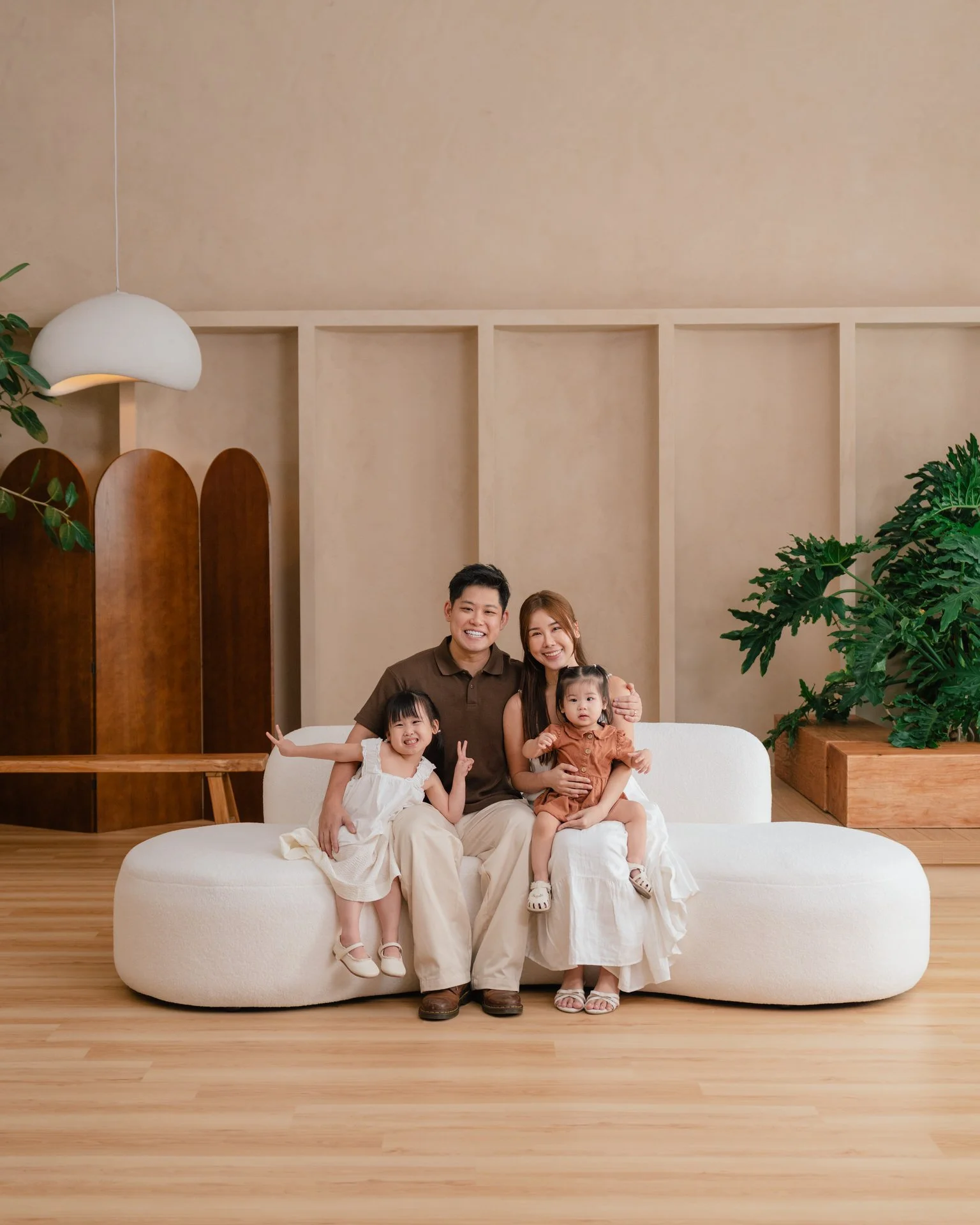 A group of eight people, including children and adults, sitting on a white round sofa in a modern, minimalistic room with a large potted tree, wooden accents, and beige walls, all smiling and laughing.