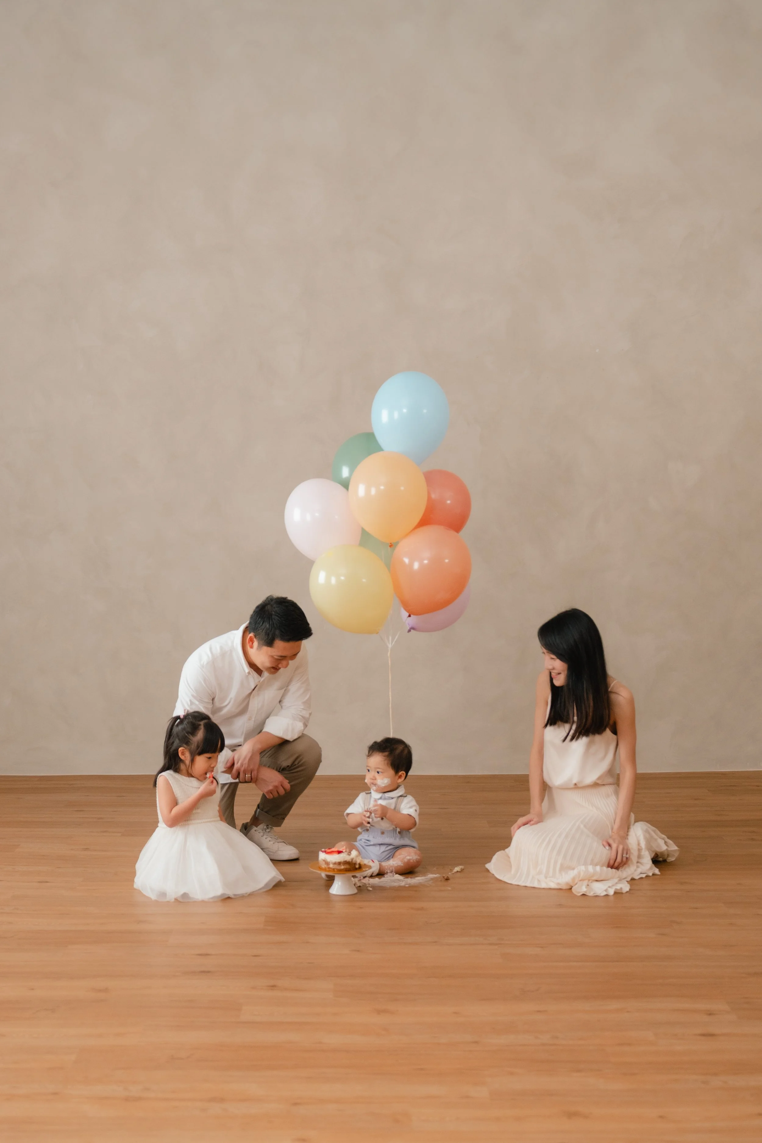 Family celebrating a child's birthday with balloons and cake in a minimal indoor setting.