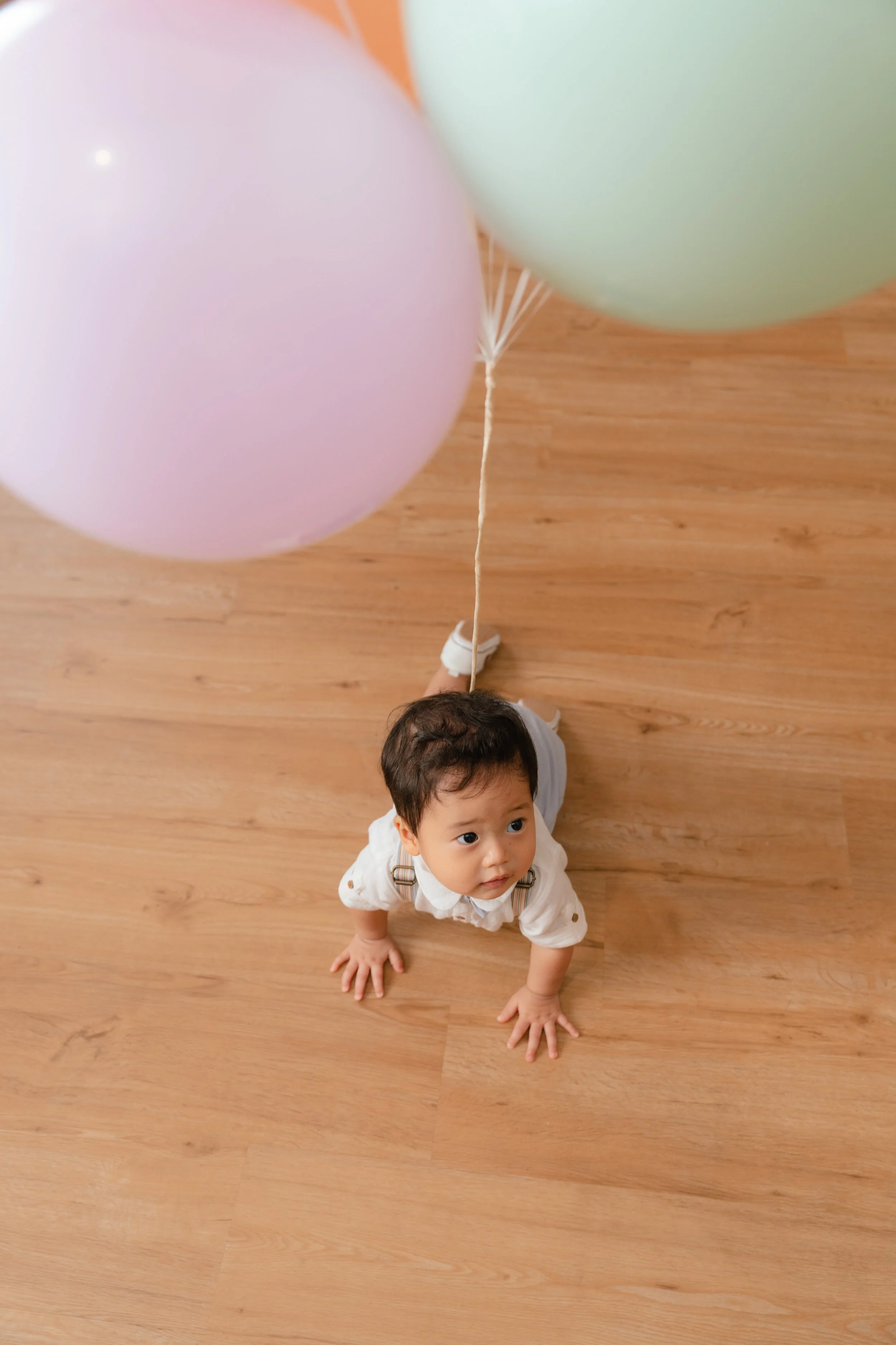 A baby looking up from the wooden floor, holding balloons tied to their waist.