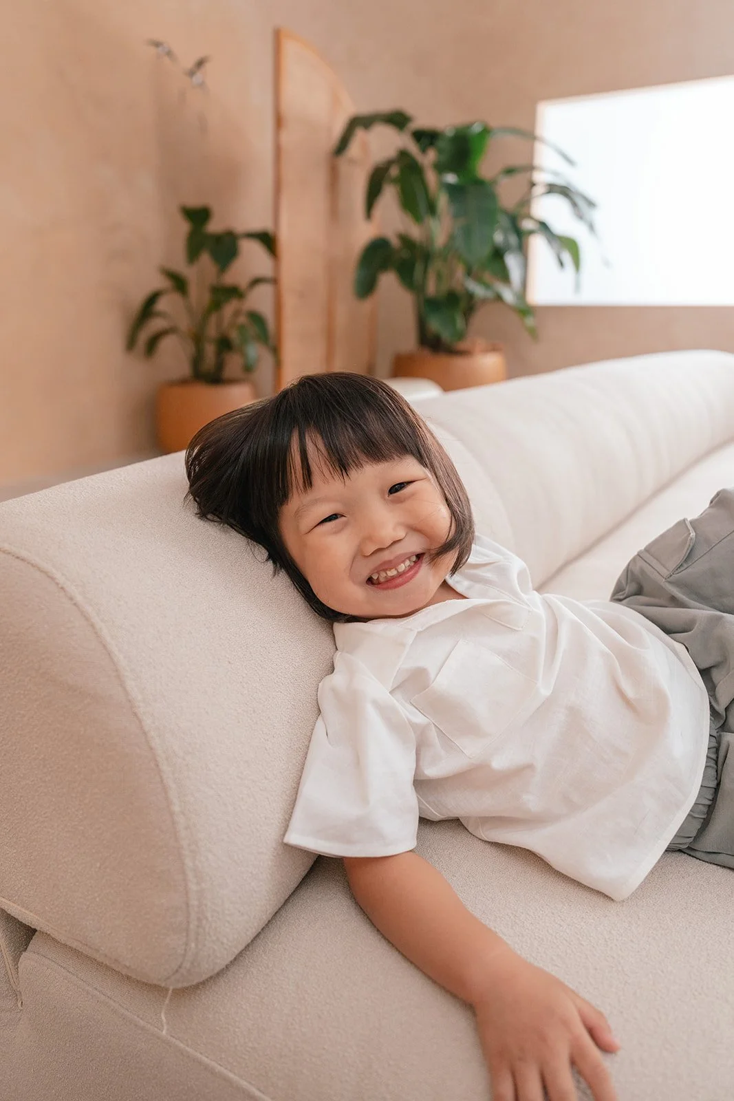 A young girl with short dark hair and a big smile laying on a cream-colored couch, wearing a white shirt and gray shorts, with potted plants and a wooden wall in the background.
