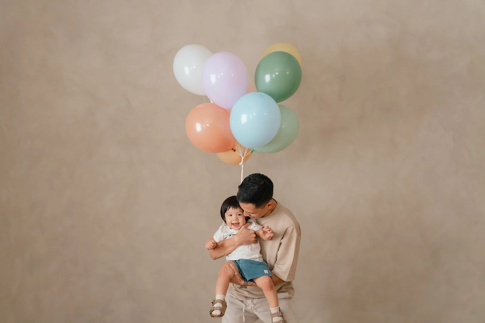 Young man holding a smiling child with colorful balloons against a beige background.