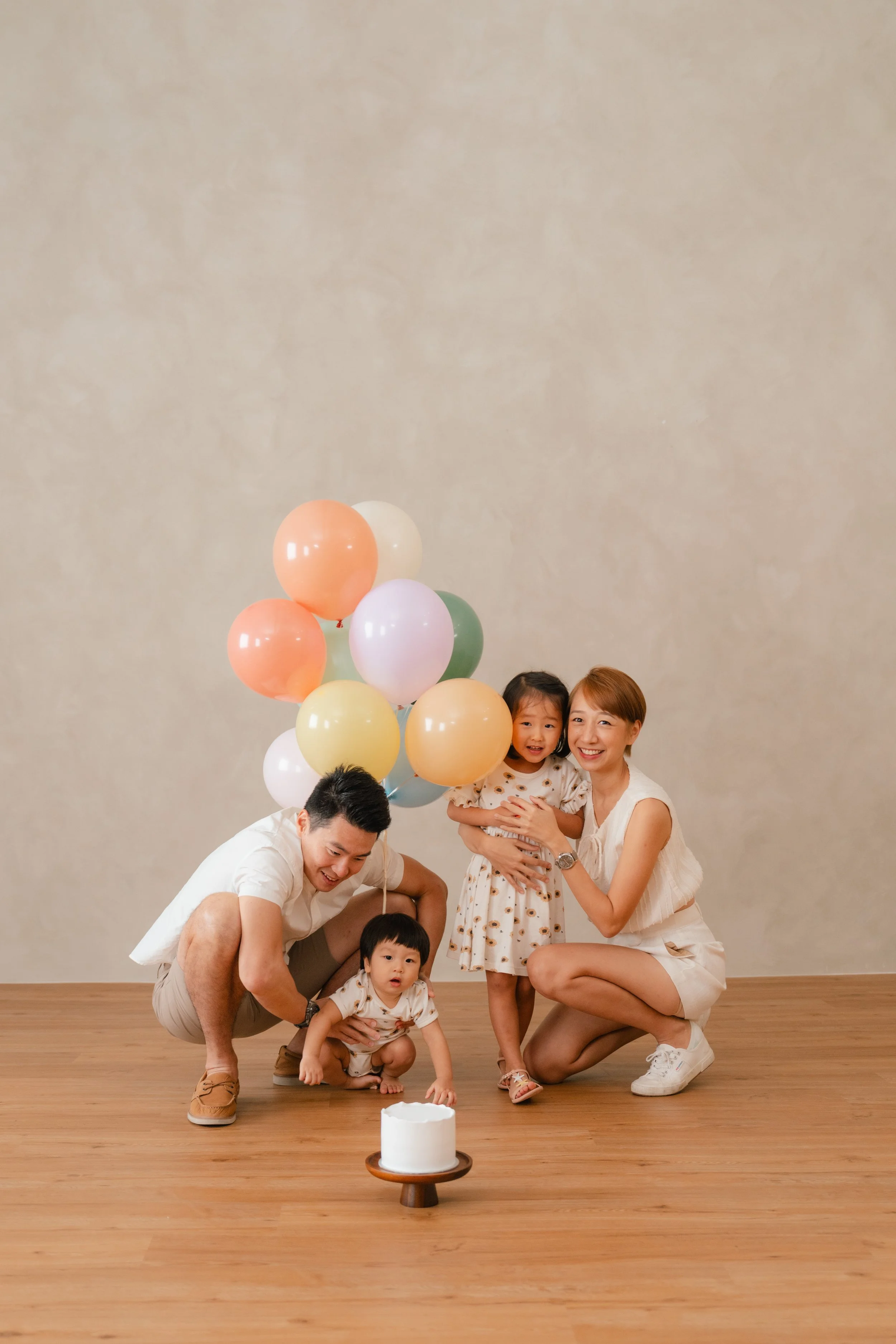 A family of four celebrating with a small cake and colorful balloons in a room with plain beige walls and wooden floor.